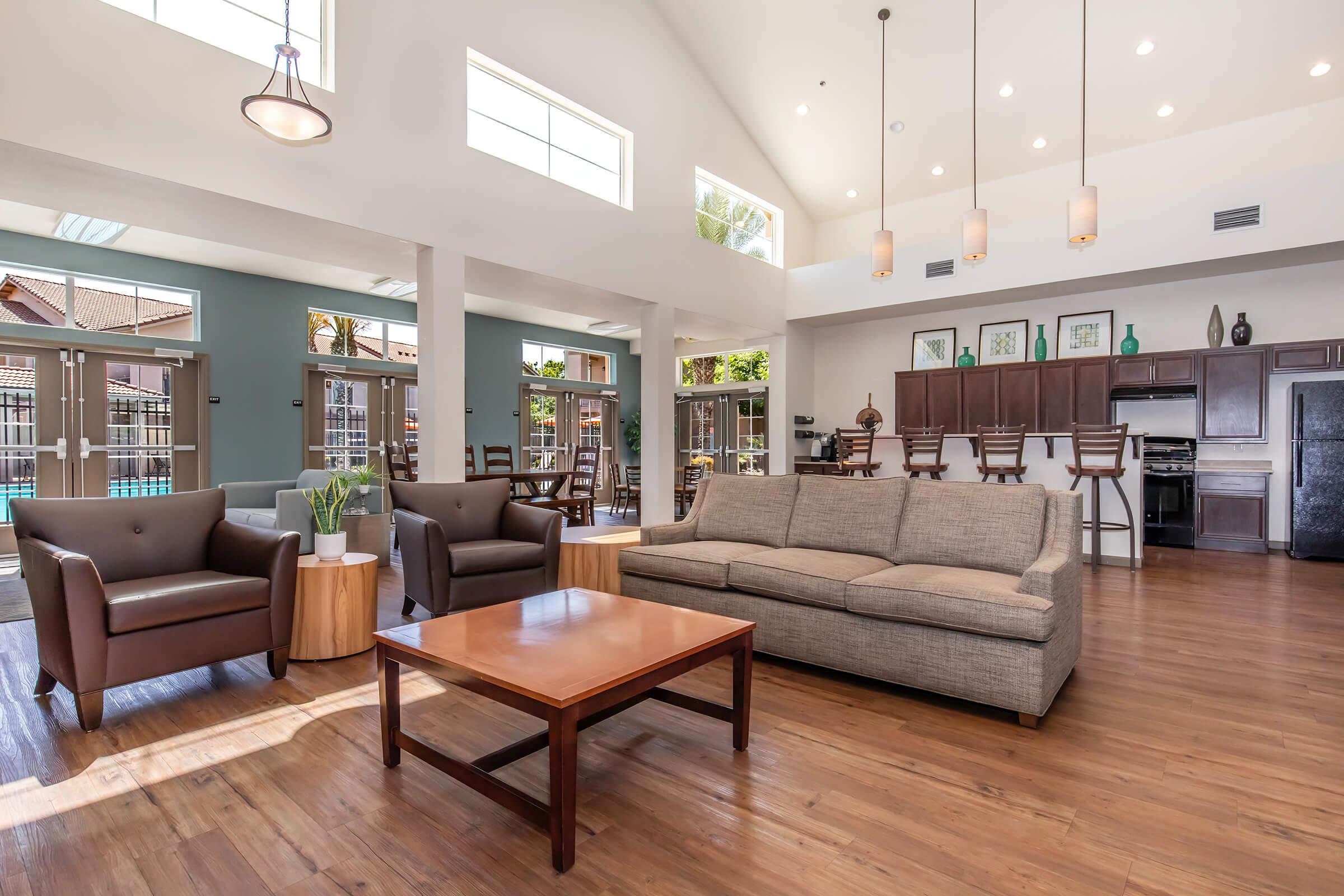 Bright and spacious living area featuring a light-colored sofa, two armchairs, and a wooden coffee table. Large windows allow natural light to fill the space, showcasing a modern design with high ceilings and stylish light fixtures. In the background, a dining area, kitchen, and pool view can be seen.