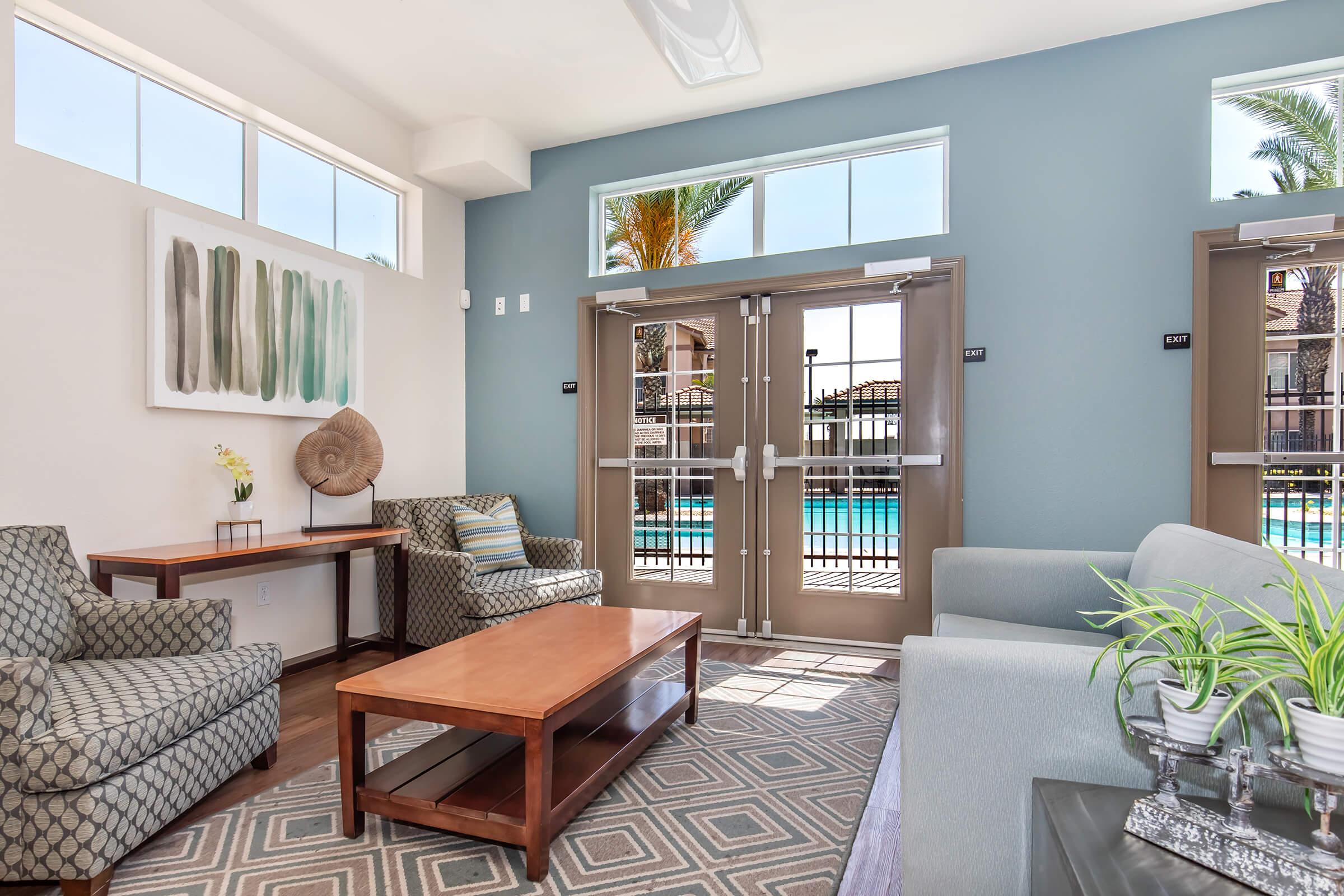 A modern lounge area featuring two patterned chairs, a wooden coffee table, and a light-colored sofa. Large windows allow natural light in, showcasing a pool area outside. Decor includes a wall art piece with green and gray tones, and potted plants add a touch of greenery. The overall ambiance is inviting and relaxed.