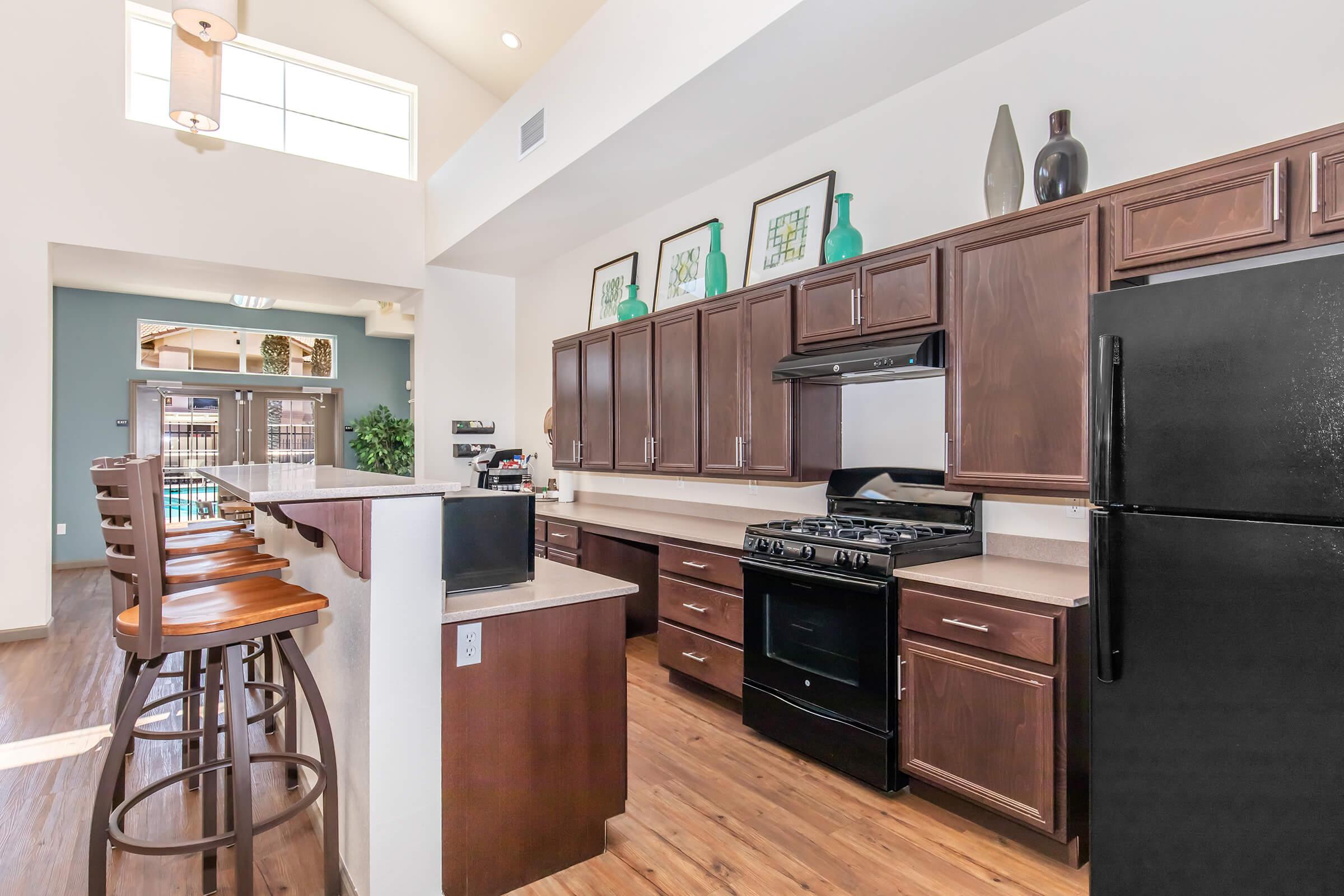 Modern kitchen featuring dark wood cabinets and a black gas stove. The countertop is beige, and the kitchen includes a bar with wooden stools. Large windows provide natural light, illuminating the space. Decorative vases and artwork are displayed on the shelves above the cabinetry.