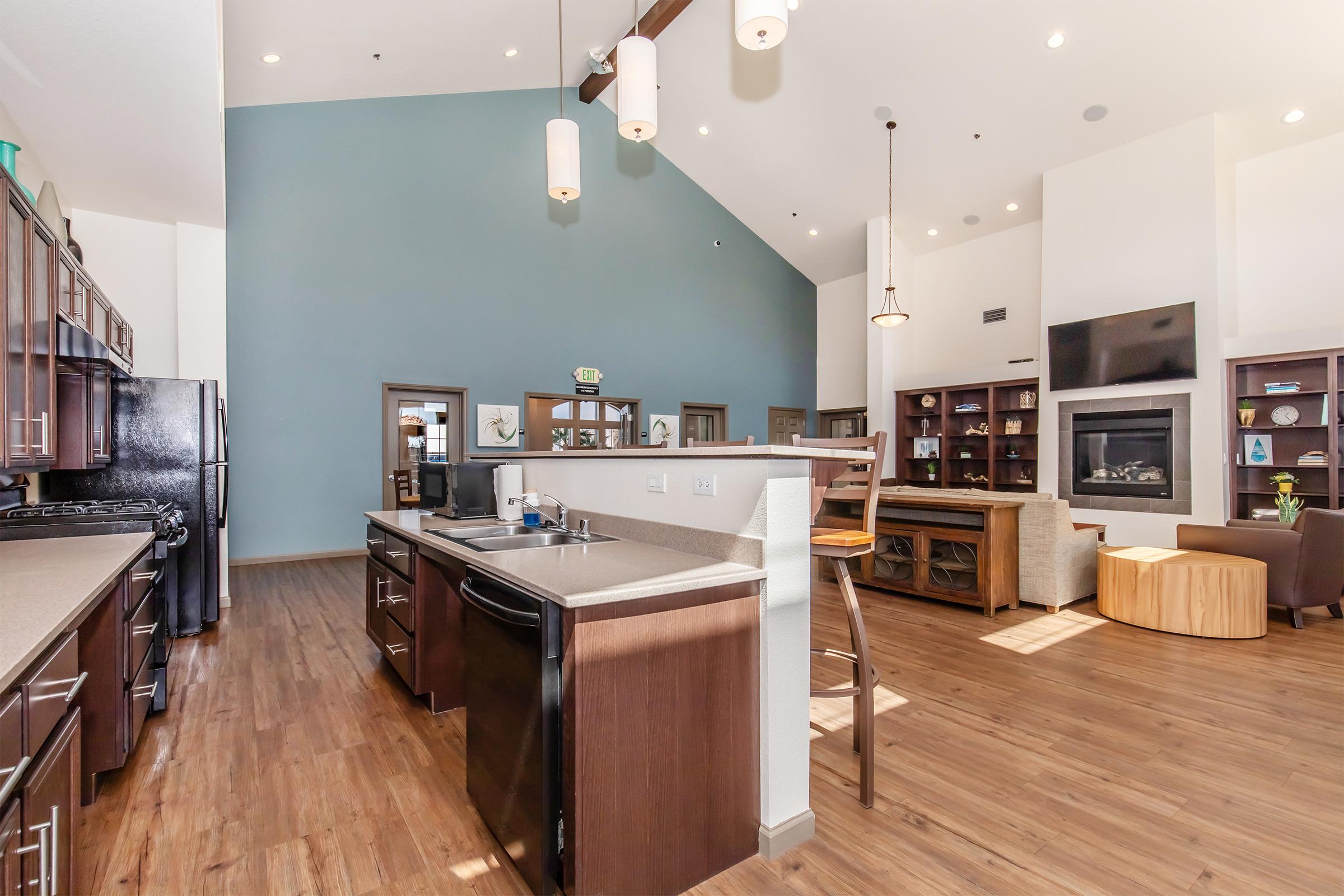 Interior view of a modern kitchen and living area featuring wooden cabinets, stainless steel appliances, a large island with seating, and a cozy seating arrangement. The walls are painted in neutral tones with one accent wall in blue, and natural light floods the space through large windows, creating a warm atmosphere.