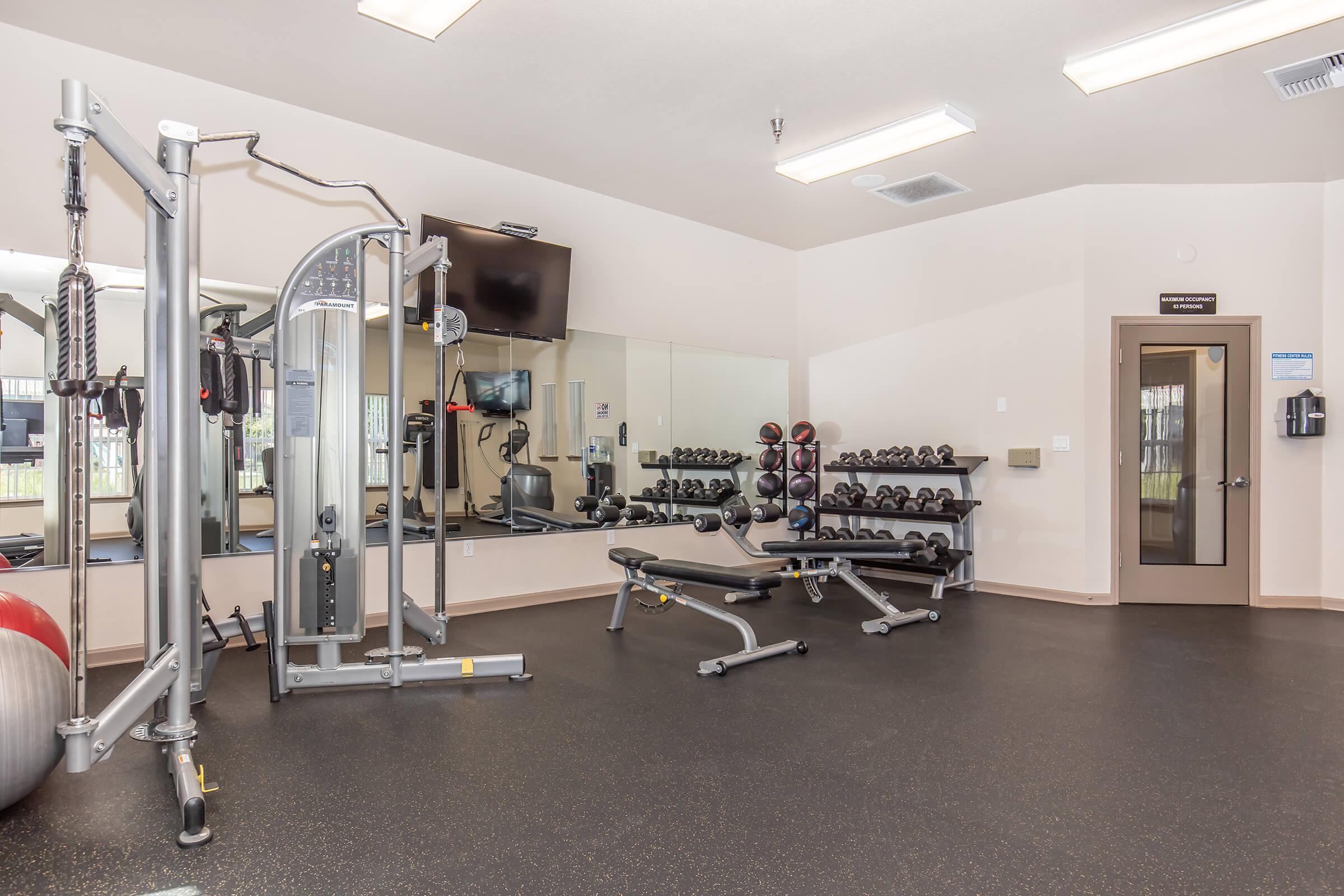 A modern gym interior featuring a multi-station weight machine, free weights organized on racks, a bench press, and mirrors on the wall. The space is well-lit with overhead lights and has exercise equipment neatly arranged, providing a clean and functional workout environment.