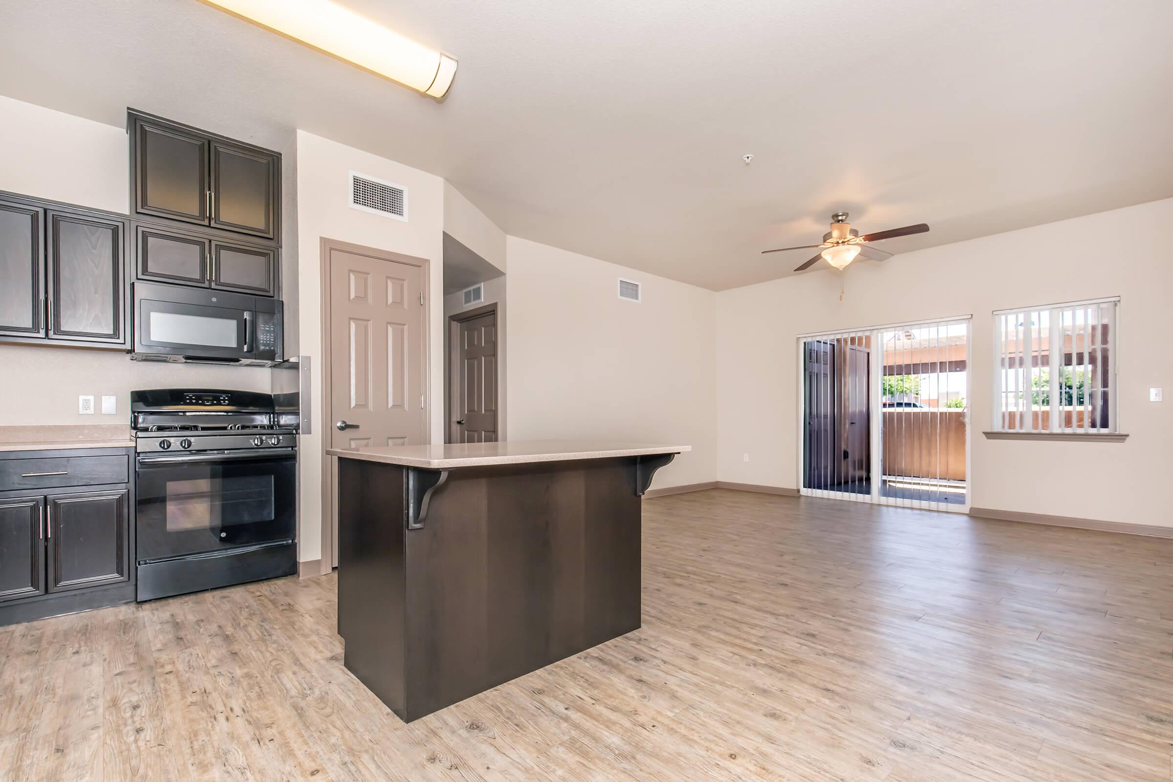 Modern open-concept living space featuring a kitchen with dark cabinetry and black appliances. A central island provides additional counter space, and large windows offer natural light. The room has wood-like flooring and leads to an outdoor area with sliding doors. Ceiling fan visible in the living area.
