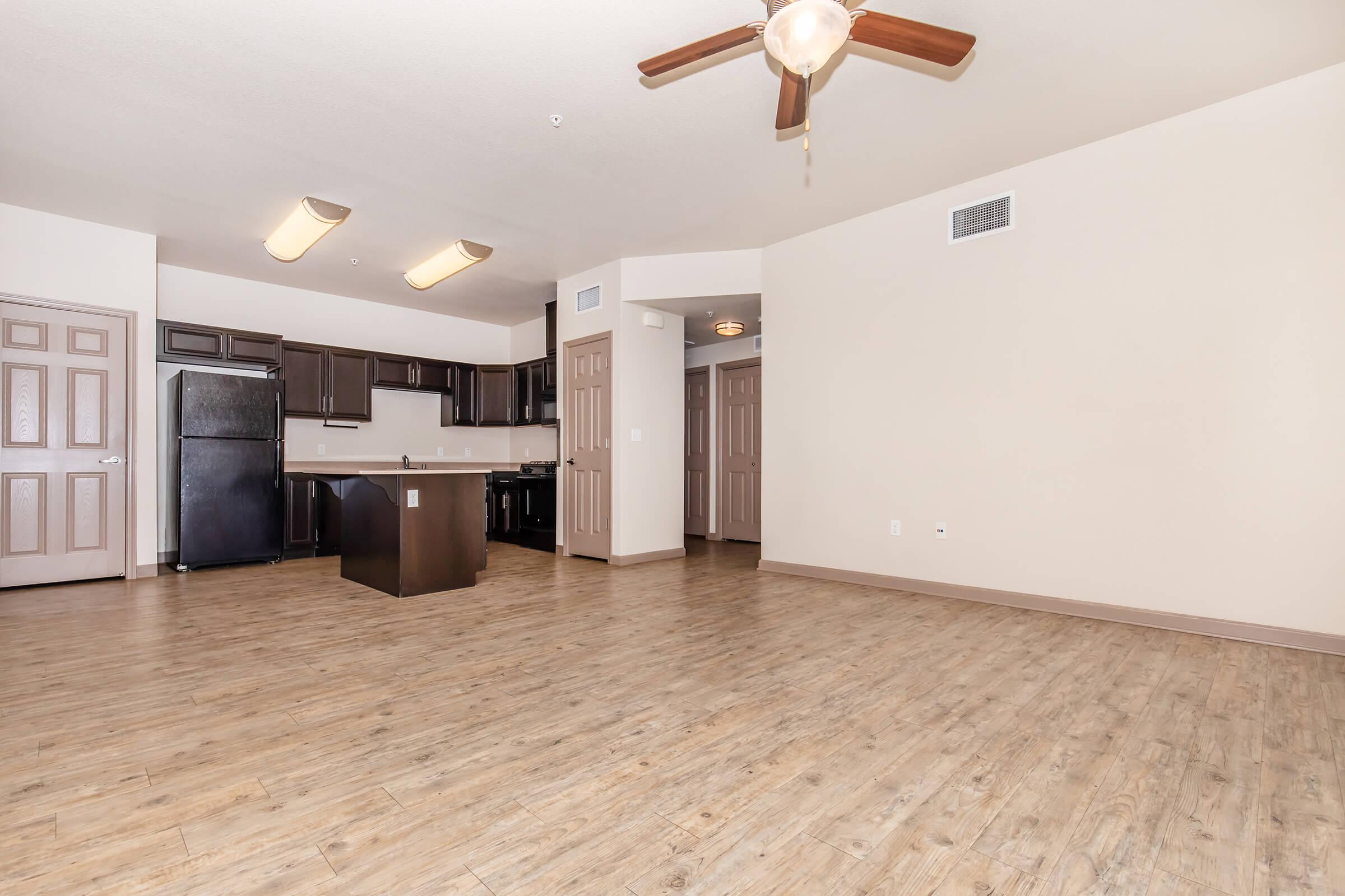 Modern open-concept living space featuring hardwood-style flooring, a ceiling fan, and kitchen with dark cabinetry and an island. There are doorways leading to other rooms, and ample natural light illuminating the area. Neutral-colored walls create a spacious and inviting atmosphere.