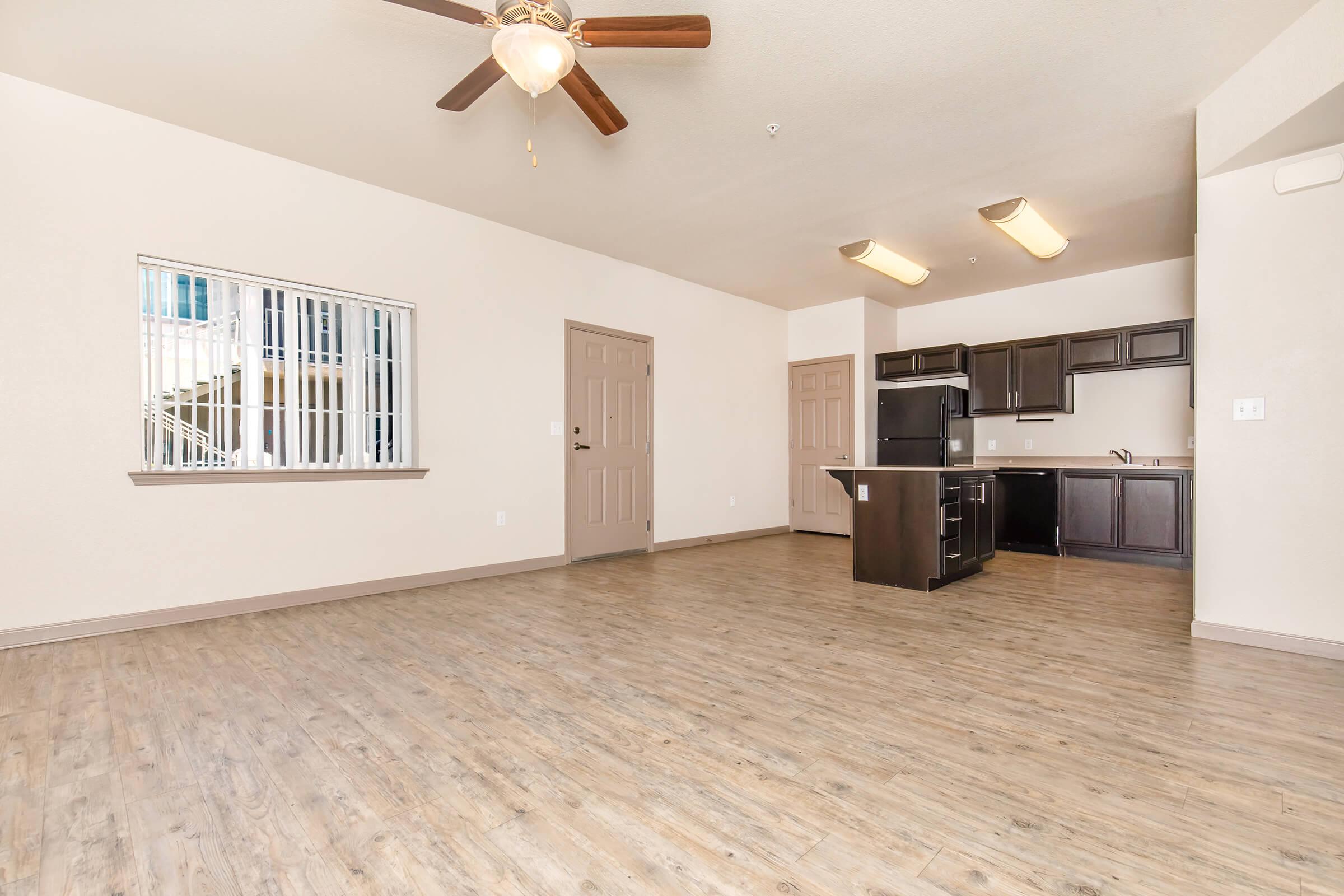 Spacious living area with wooden floors, a ceiling fan, and a window. The kitchen area features dark cabinets and appliances, visible in the background. The walls are painted in a light neutral color, and there are two doors leading to other rooms.