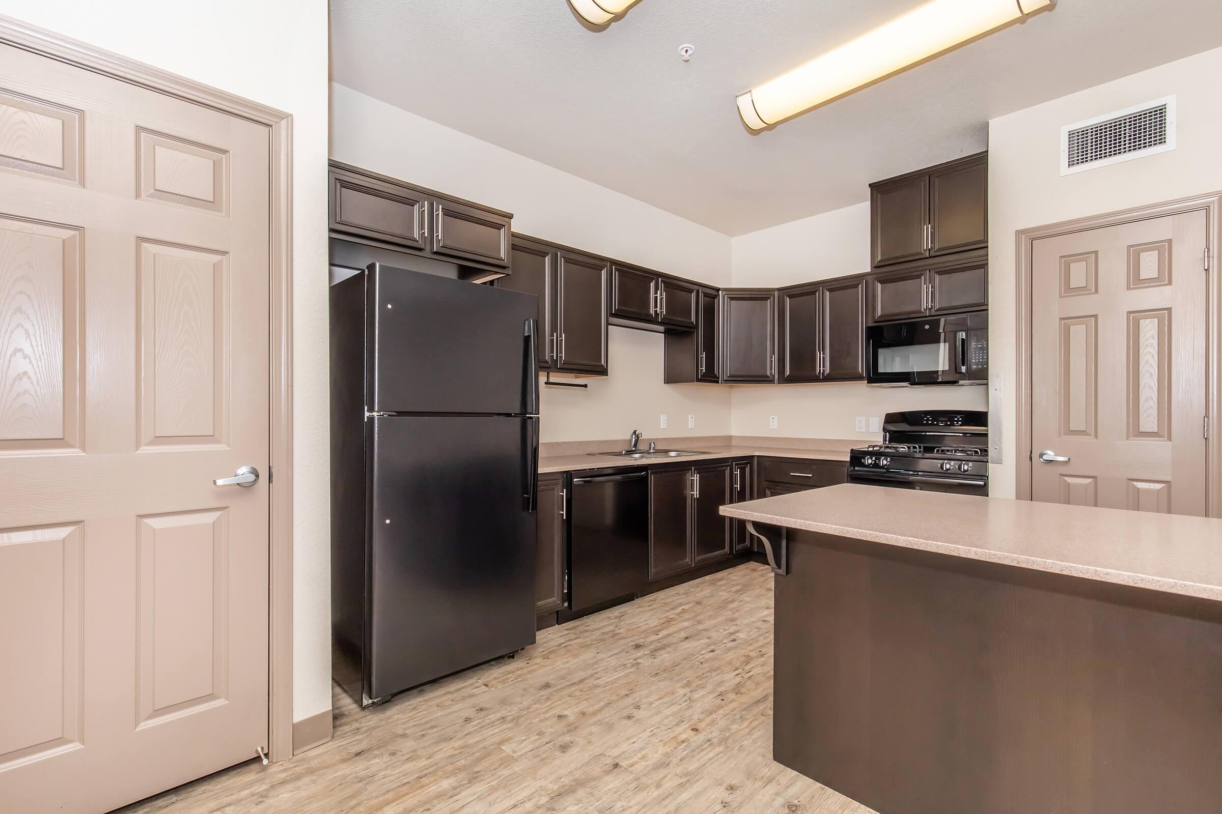 Modern kitchen featuring dark wood cabinets, black appliances including a refrigerator and stove, and a spacious countertop. The floor has a wood-like finish, and the walls are painted in a neutral color. Ample cabinet space and a microwave are also included, creating a stylish and functional cooking area.