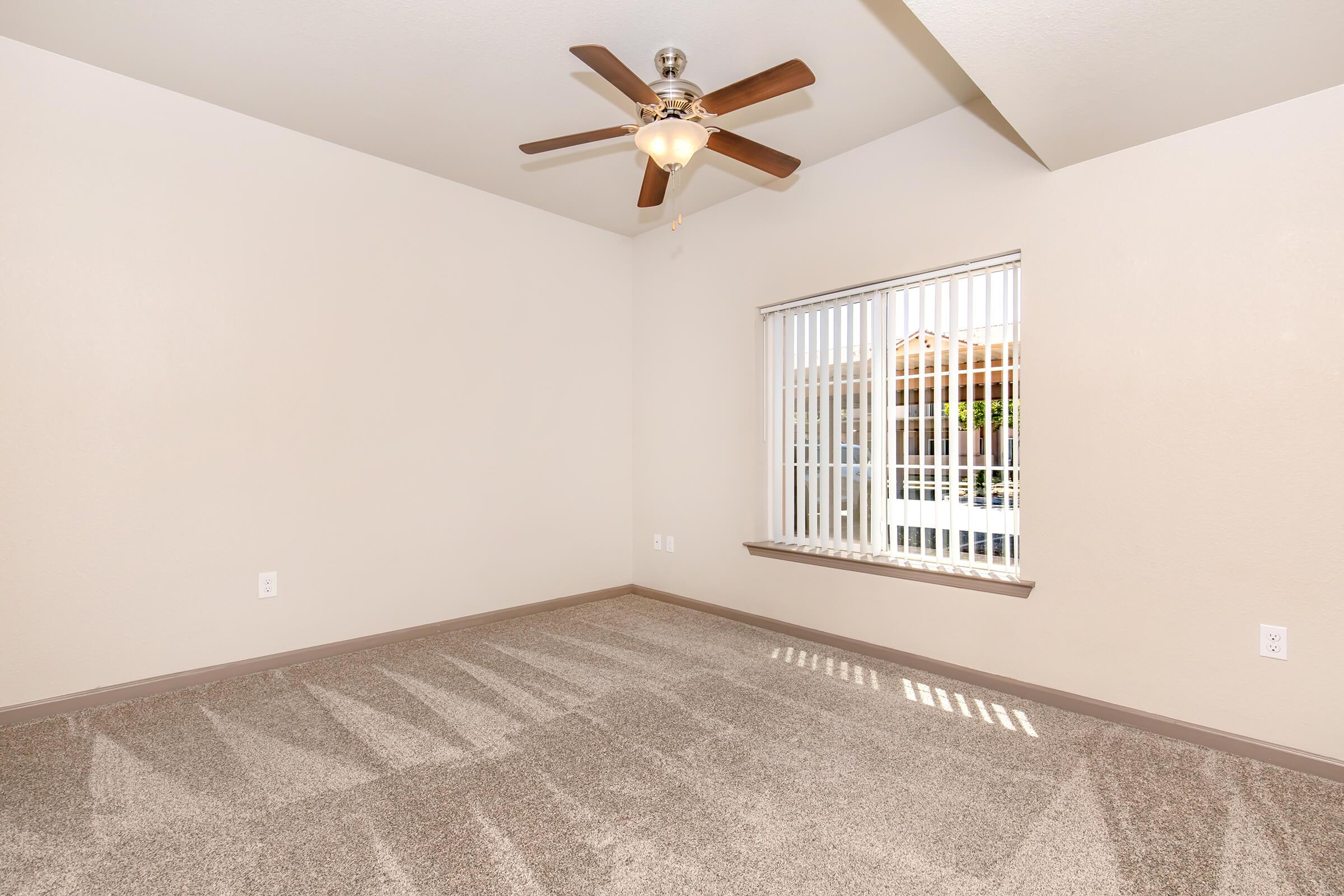 A well-lit room featuring beige walls and a ceiling fan with wooden blades. The space has soft carpeting and a window with vertical blinds allowing natural light in. The room is empty, showcasing its spaciousness, making it suitable for various furnishings.