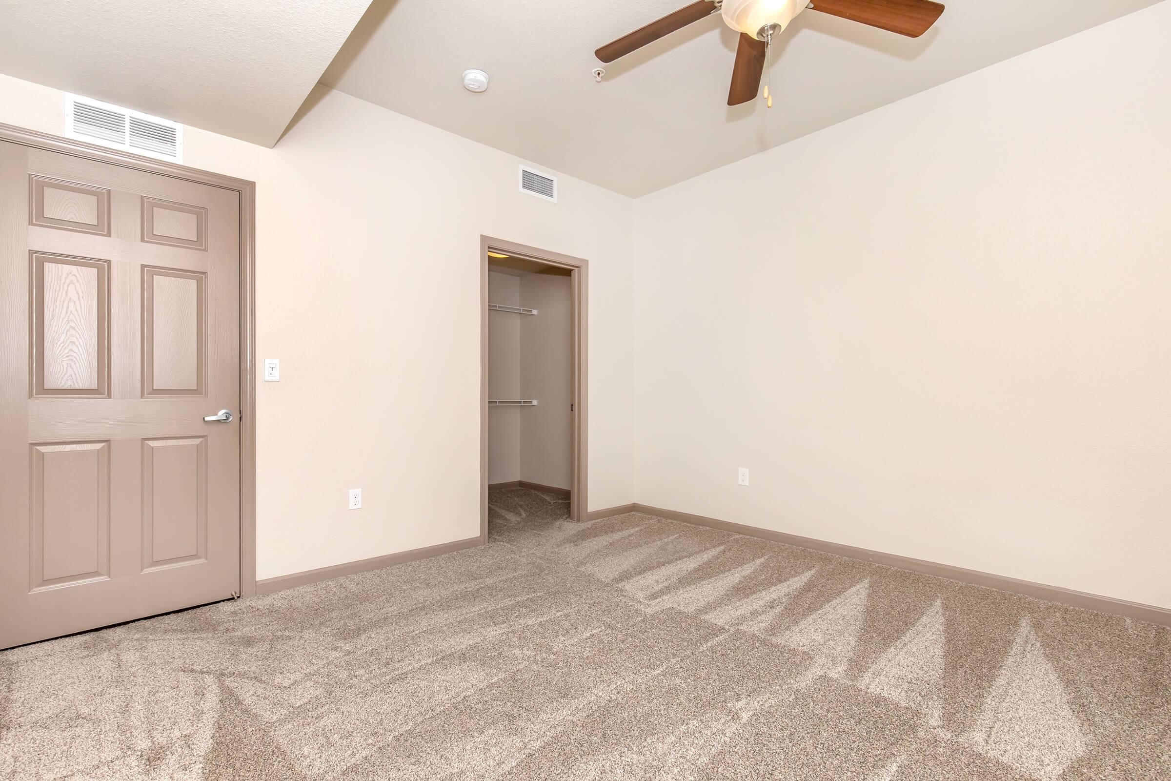 A small, minimalistic room featuring beige walls and carpet, a ceiling fan, and a closed door leading to another space. There is a closet with an open door on the left side. Natural light creates shadow patterns on the floor, enhancing the simple and clean aesthetic of the room.