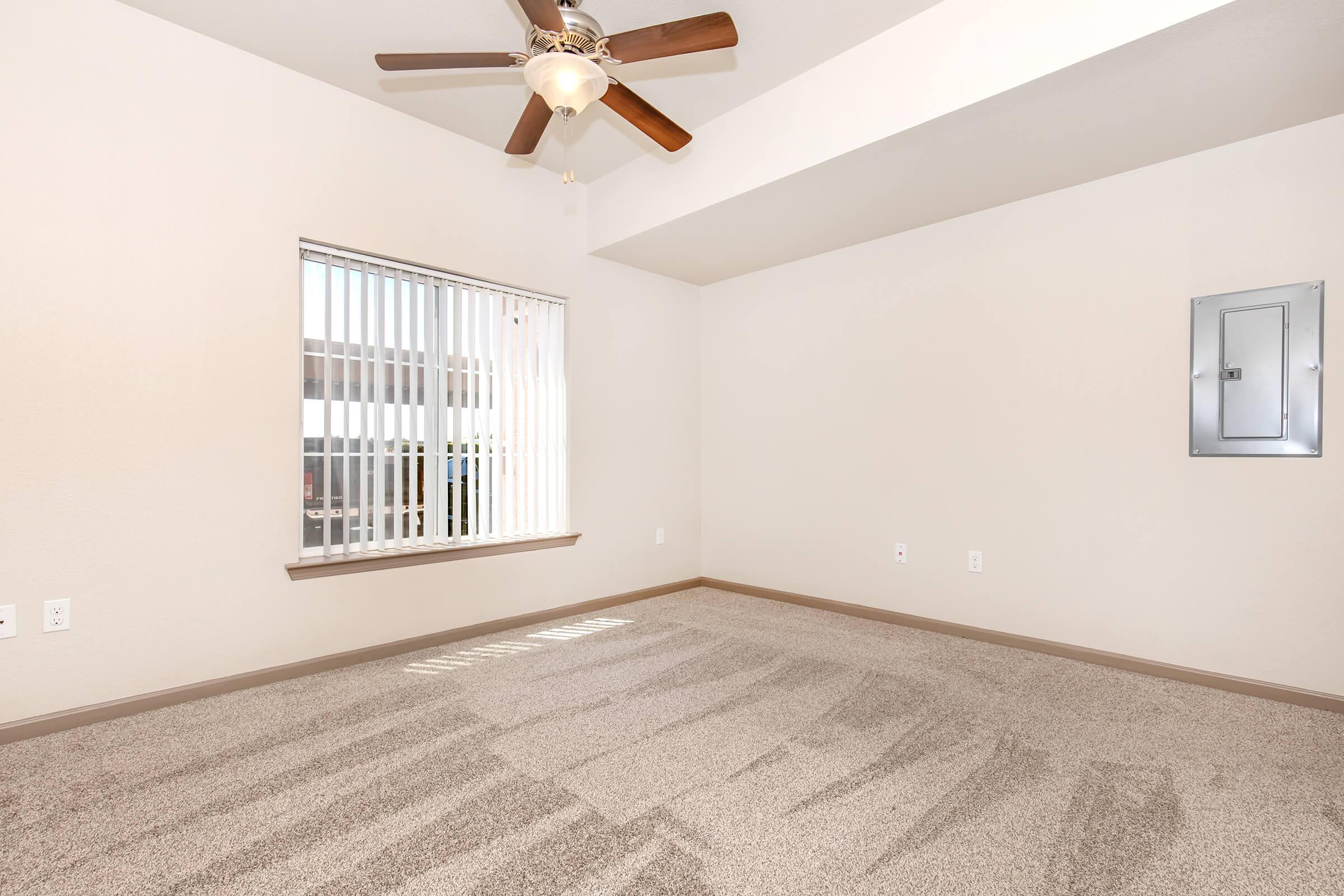 A spacious, empty room with light beige walls and a ceiling fan. Sunlight streams through a window with vertical blinds, casting shadows on the plush, light gray carpet. There is a small electrical panel on one wall, contributing to a clean and minimalist design.