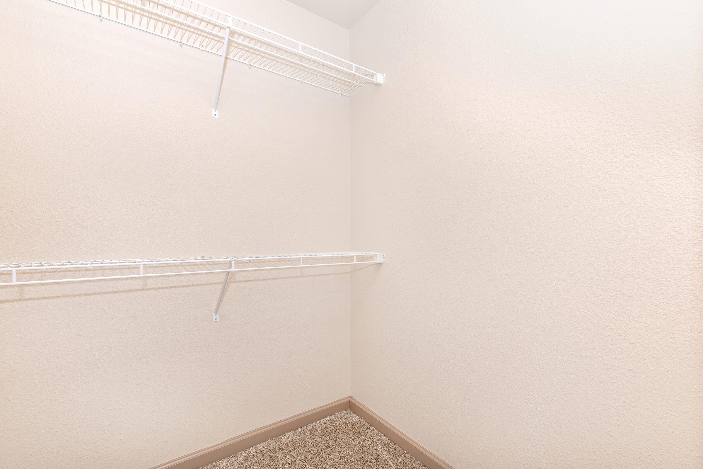 Empty closet space with light beige walls and carpeted floor. There are two wire shelves mounted on the wall, one on the left side and one on the back wall, creating a minimalist storage area. The overall appearance is clean and uncluttered.