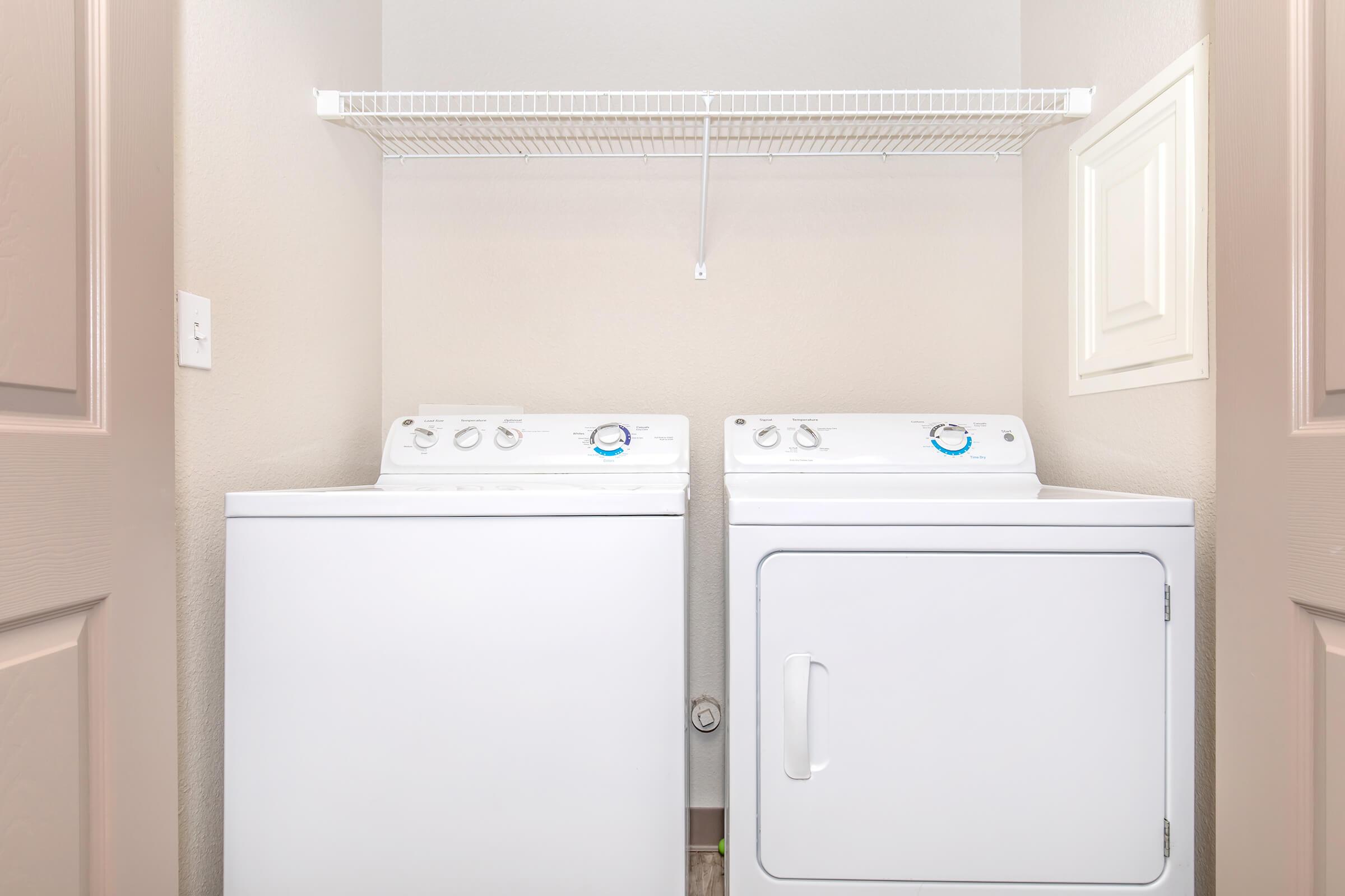 A white washing machine and a white dryer are positioned side by side in a small laundry room. Above them, a wire shelf is mounted on the wall, providing storage space. The walls are a light color, contributing to a clean, organized appearance.
