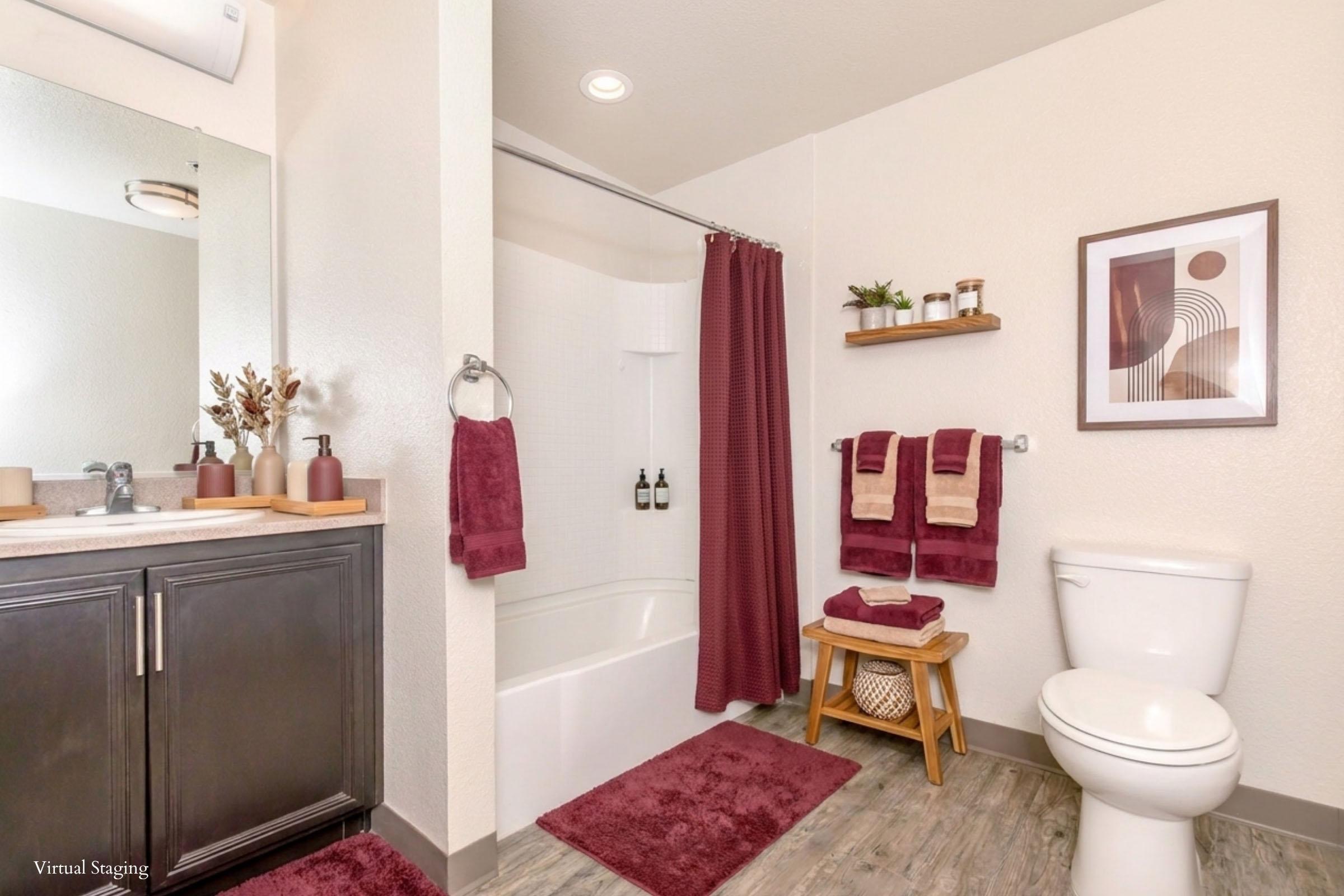 A clean and modern bathroom featuring a white bathtub with a shower curtain, a wooden vanity with a mirror, and a toilet. The space is decorated with maroon towels and accessories, a small wooden stool, and a piece of wall art, creating a cozy and inviting atmosphere.