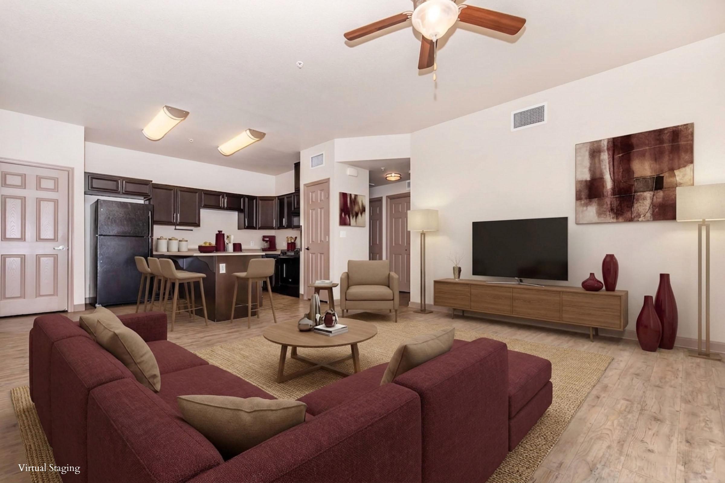 A modern living room featuring a burgundy sofa set, a wooden coffee table, and a stylish TV stand with a large flat-screen TV. In the background, there is a kitchen area with dark cabinetry. The space is well-lit with ceiling lights and a floor lamp, and neutral walls provide a warm ambiance. A decorative rug adds comfort to the room.