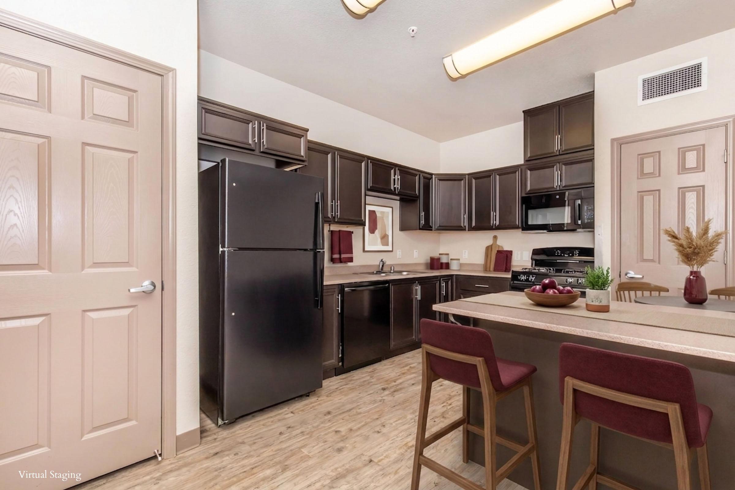 Modern kitchen featuring dark cabinetry, stainless steel appliances, and a large island with seating. The space is well-lit, showcasing a blend of functionality and style, with decorative accents such as a framed artwork and a small plant. Warm wooden floors complement the contemporary design.