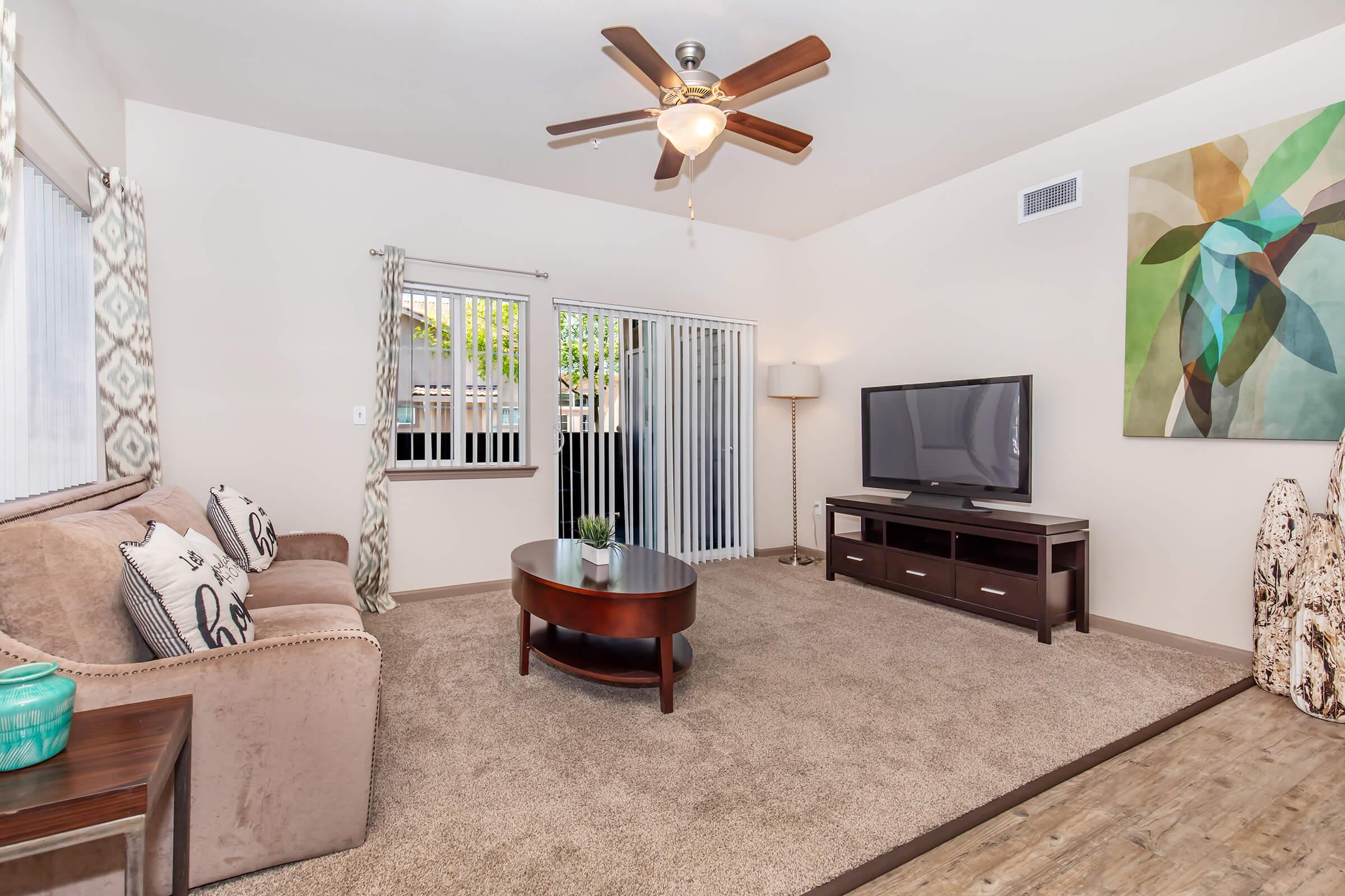 A cozy living room featuring a beige sofa with decorative pillows, a coffee table, and a dark wood TV stand with a flat-screen TV. There are large windows with sheer curtains and a sliding door, allowing natural light. A colorful abstract painting hangs on the wall, enhancing the modern decor.
