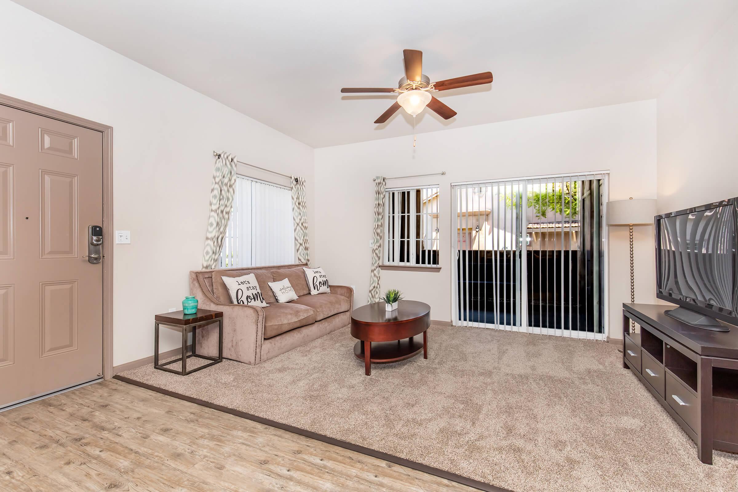 A cozy living room featuring a brown sofa with decorative pillows, a small round coffee table, and a television on an entertainment unit. Natural light streams in through vertical blinds on large windows. The room has a light-colored wall, ceiling fan, and carpeted flooring, creating a warm and inviting atmosphere.