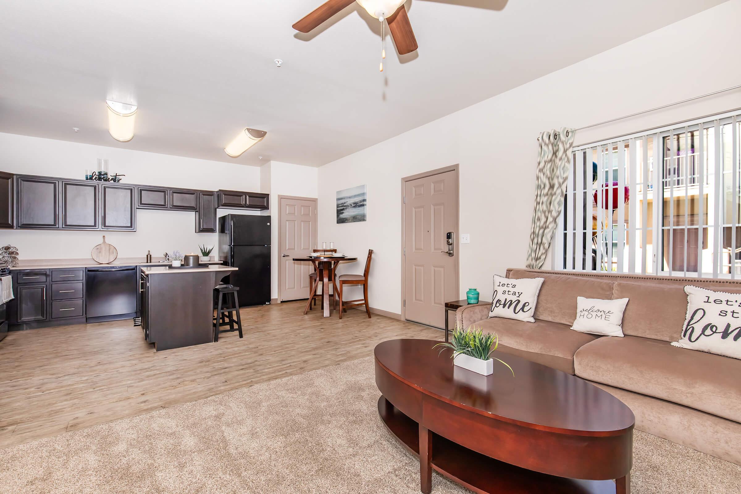 A cozy living space featuring a living room with a brown couch and round coffee table, a kitchen with dark cabinets and appliances, a dining area with high chairs, and natural light filtering through window blinds. Decorative pillows on the couch convey a welcoming message.