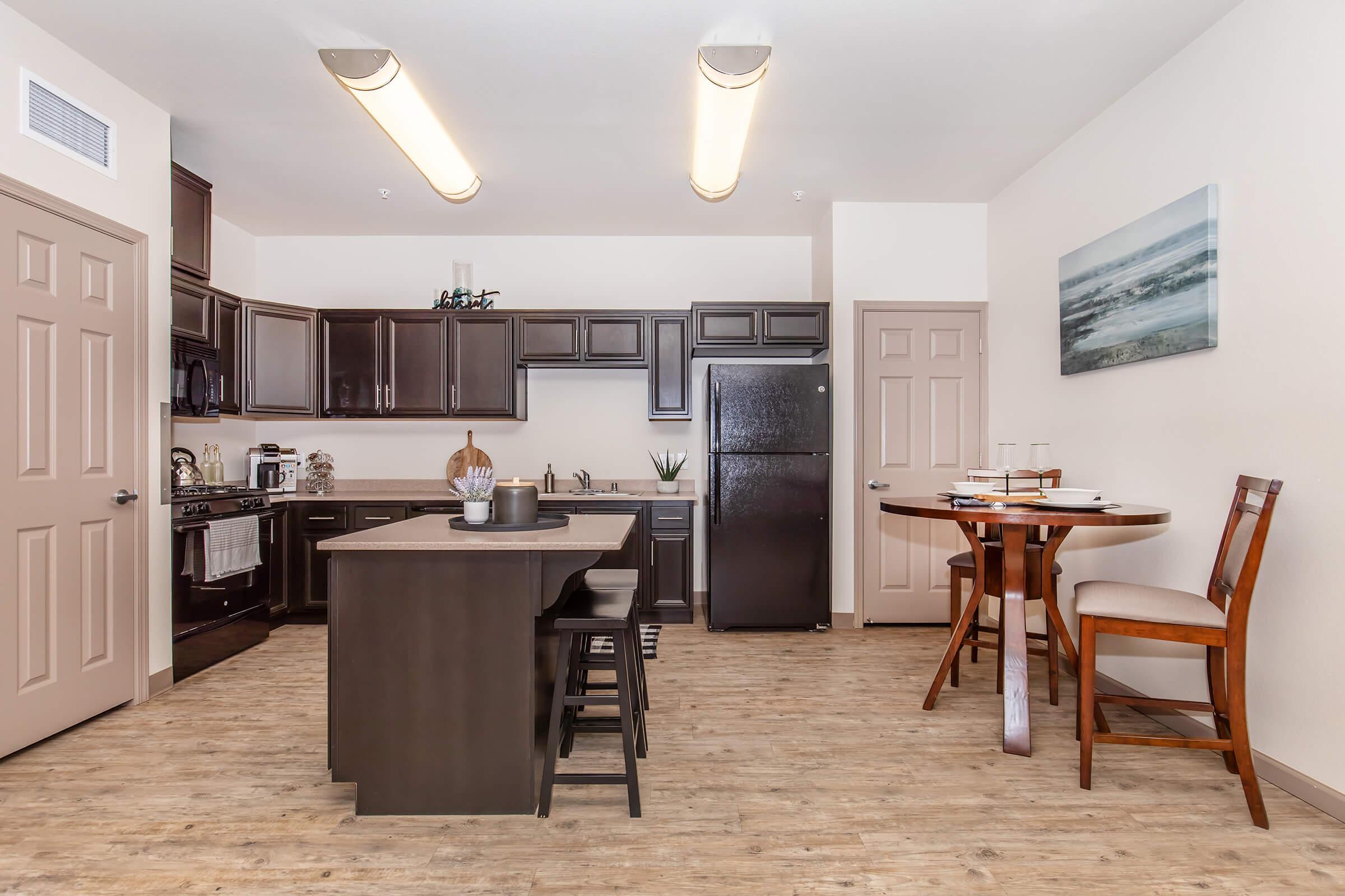 Modern kitchen and dining area featuring dark wooden cabinets, stainless steel appliances, a black refrigerator, and a round wooden table with two chairs. The floor has a light wood finish, and a large window provides natural light, enhancing the open layout and contemporary style of the space.