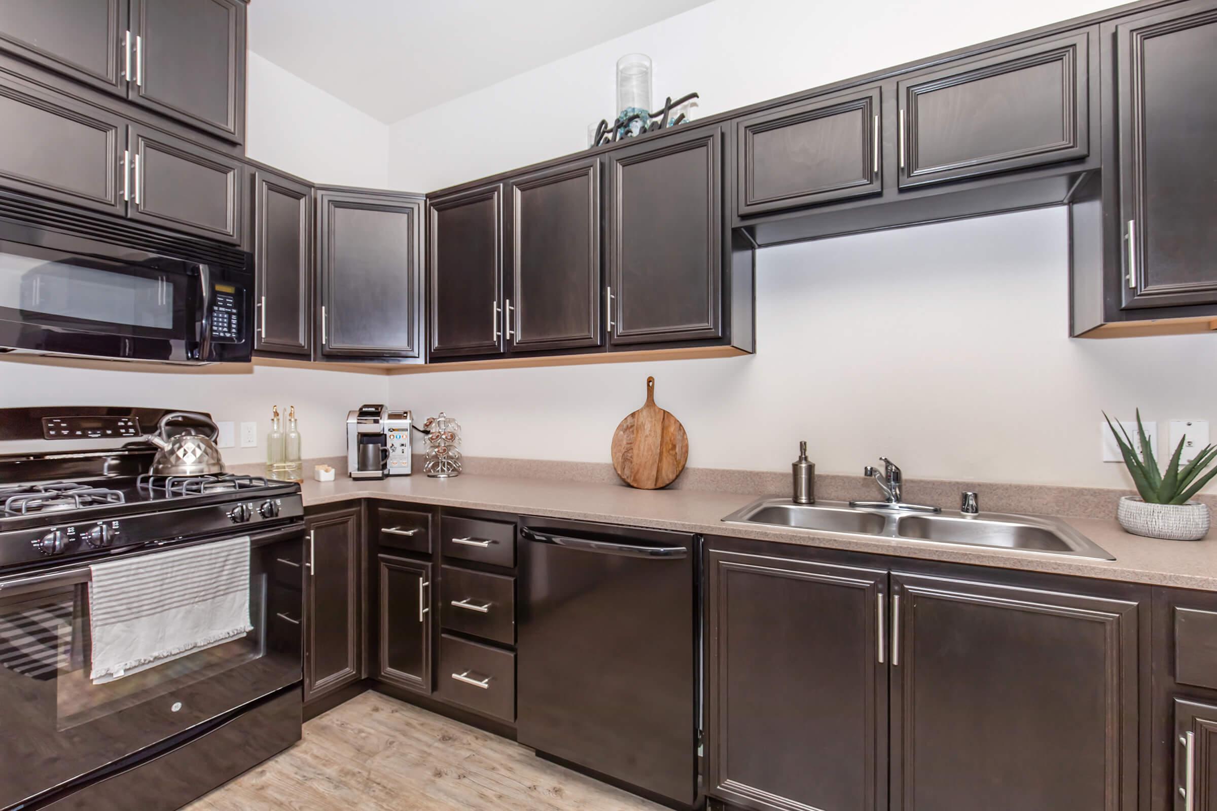 A modern kitchen featuring dark wooden cabinets, a black stove, and a microwave. The countertop is set with a cutting board, a sink, and a decorative plant. Appliances include a dishwasher and a coffee maker, with a clean, organized layout and wooden flooring.