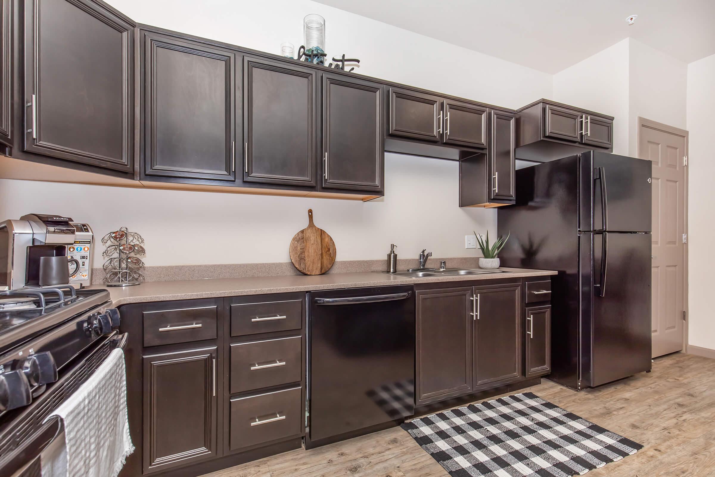 Modern kitchen featuring dark wood cabinetry, a black refrigerator, and a stainless-steel sink. There's a decorative wooden cutting board on the countertop, a small plant, and a checkered rug on the floor. The kitchen has an overall sleek and contemporary look.