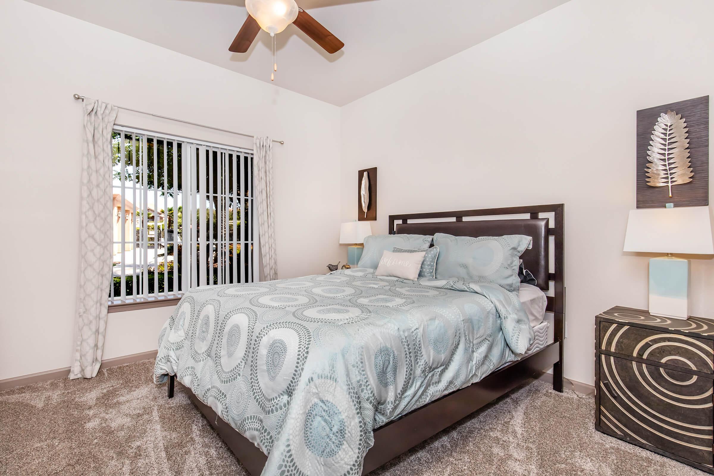 A cozy bedroom featuring a hardwood bed with a light blue patterned comforter. There are two bedside lamps and a decorative nightstand. The room has a large window with blinds, allowing natural light, and the carpeted floor adds warmth to the space.