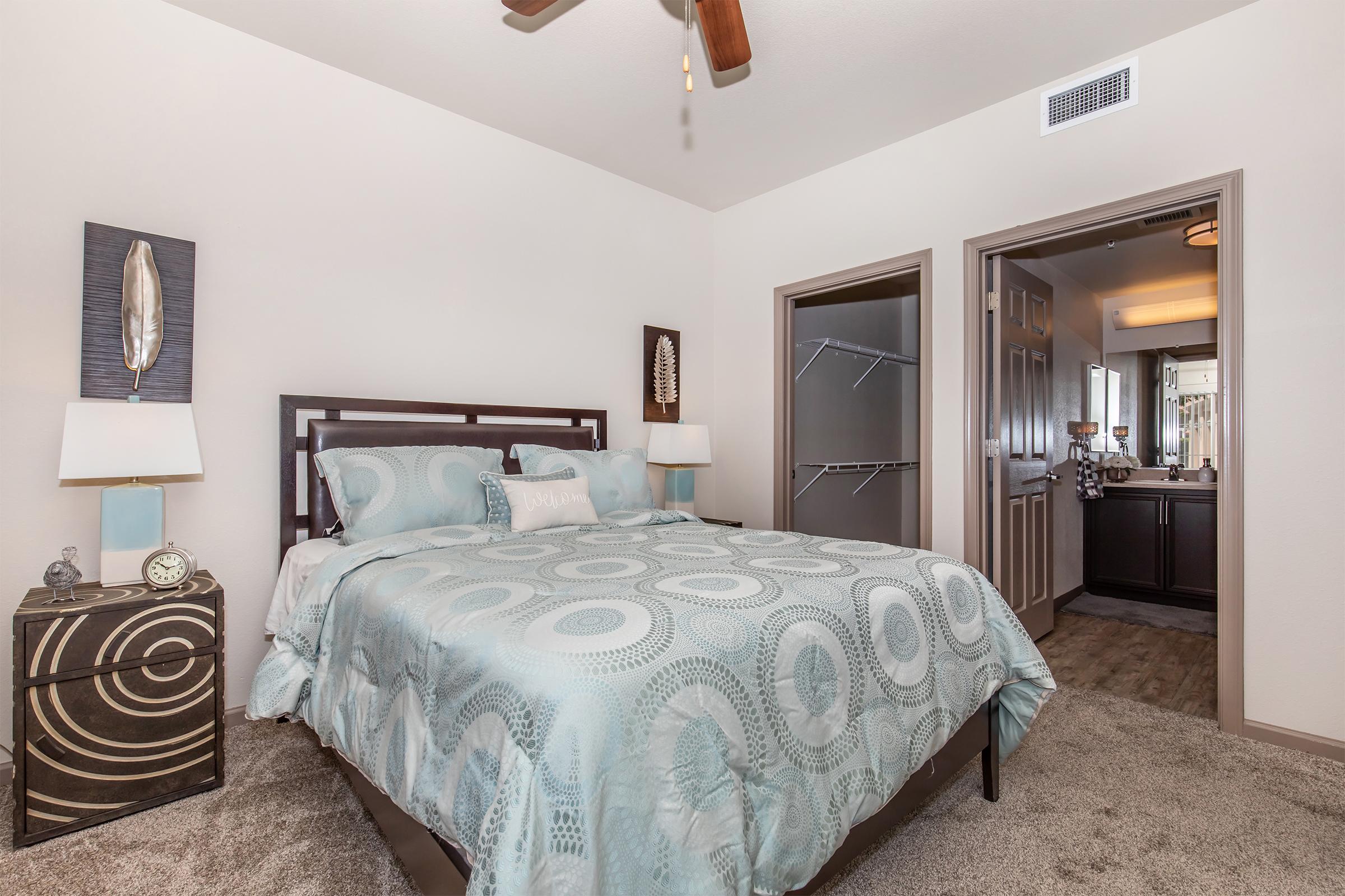A well-decorated bedroom featuring a queen-sized bed with a light blue and circular patterned comforter. There's a nightstand with a small clock and decorative item, a closet with a sliding door, and an adjoining bathroom visible through an open door. The walls are light-colored, and a ceiling fan is present.