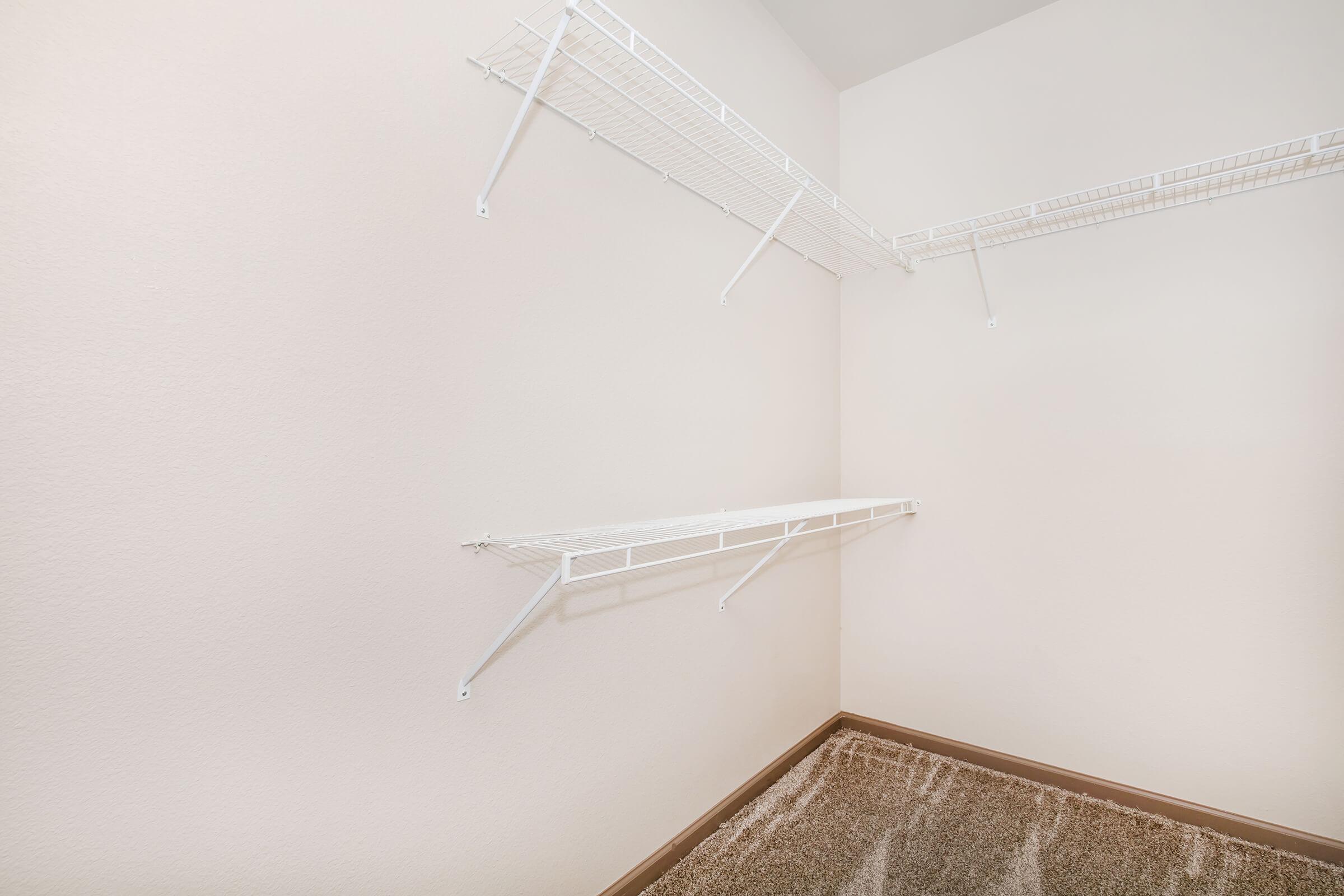 A corner of an empty closet featuring white wire shelves mounted on light-colored walls. The floor is carpeted in beige, and there are no visible items or decorations, giving a clean and spacious appearance.