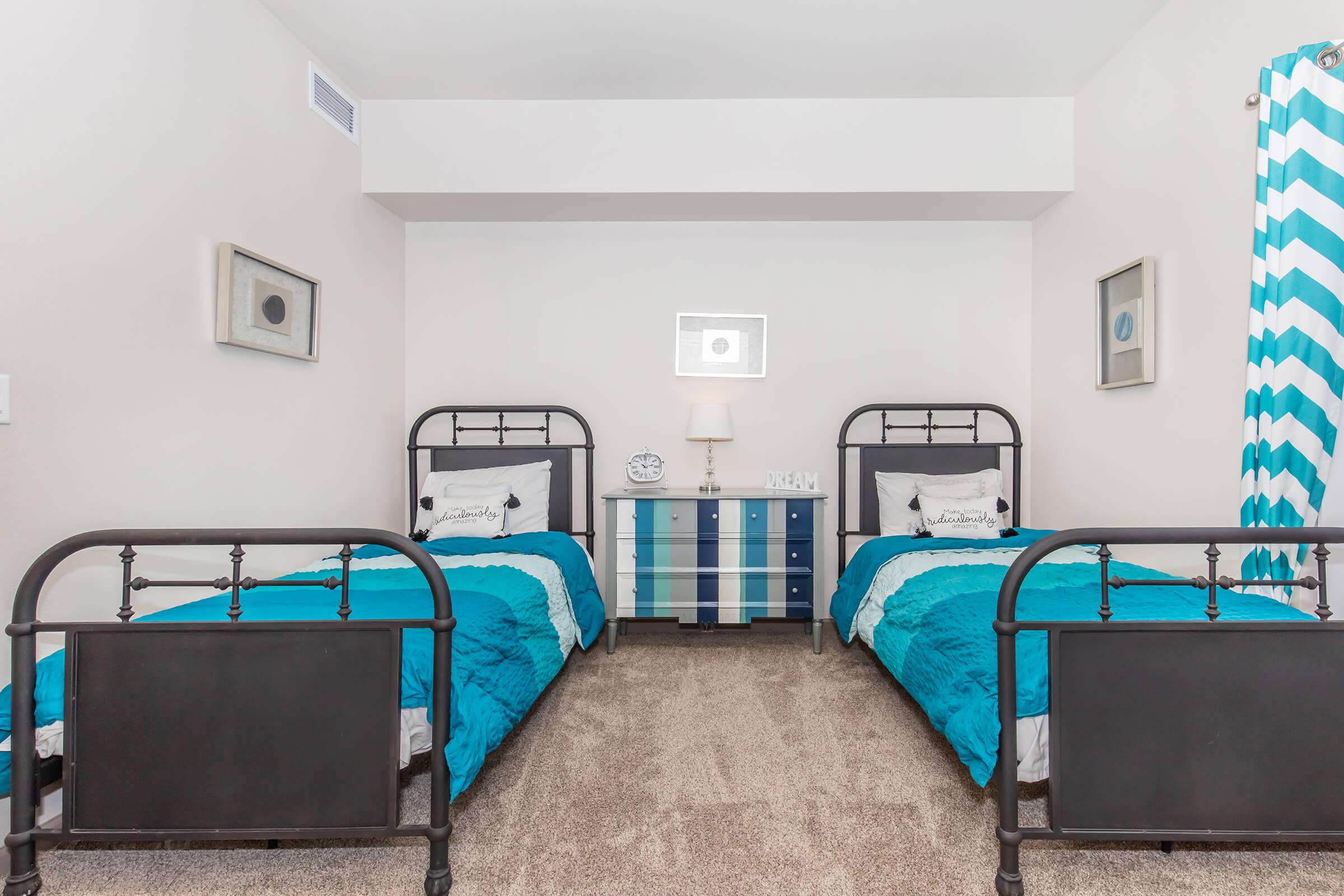 A bedroom featuring two black metal frame twin beds with bright turquoise bedding. A small nightstand with a lamp is between the beds, and a dresser with blue and white striped accents is against the wall. Light-colored walls and carpet create a modern, inviting atmosphere.
