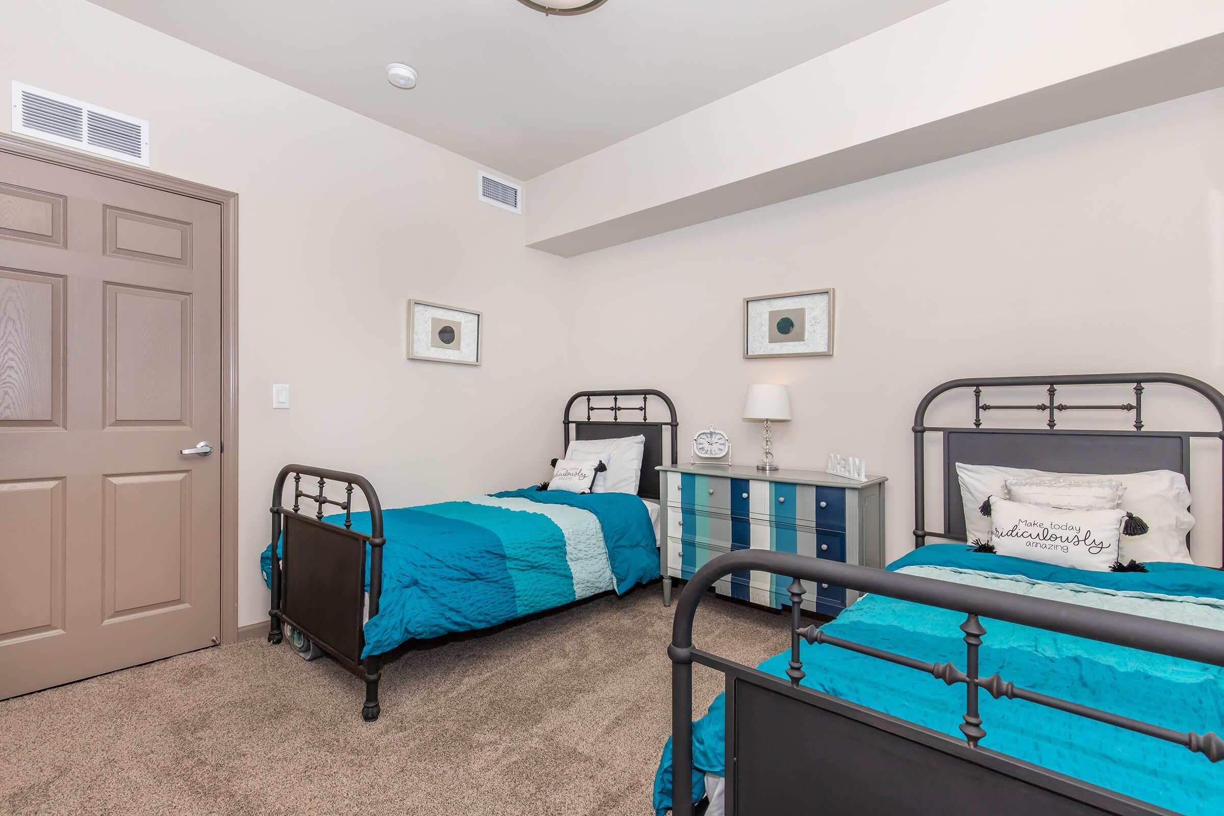A cozy bedroom featuring two twin beds with turquoise and white bedding. There's a blue dresser and framed artwork on the walls. The room has neutral beige walls and a door leading to the hallway. Soft carpet covers the floor, creating a warm and inviting atmosphere.