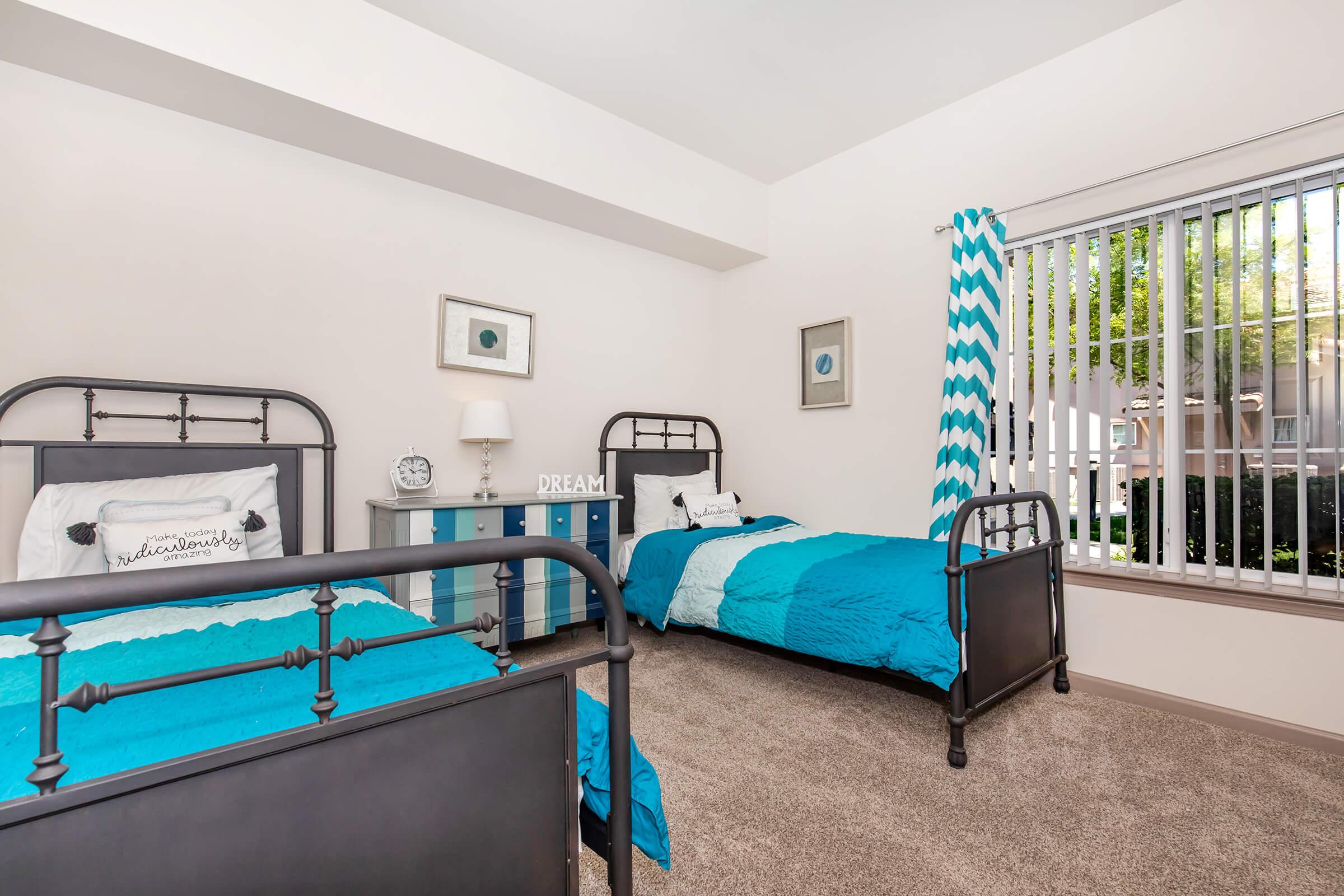 A cozy bedroom featuring two metal-frame beds dressed in turquoise-blue bedding. A nightstand with a lamp and decorative items sits between the beds. A large window with vertical blinds allows natural light, and a patterned curtain adds a touch of style. The room has a neutral wall color and plush carpet.