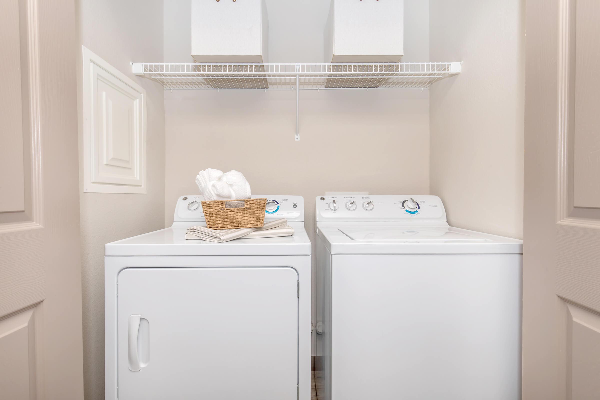A laundry room featuring a washing machine and a dryer side by side. Above them, there is a shelf holding a basket and neatly folded items. The walls are painted a light color, and the space is compact, with closed doors on the left and right.