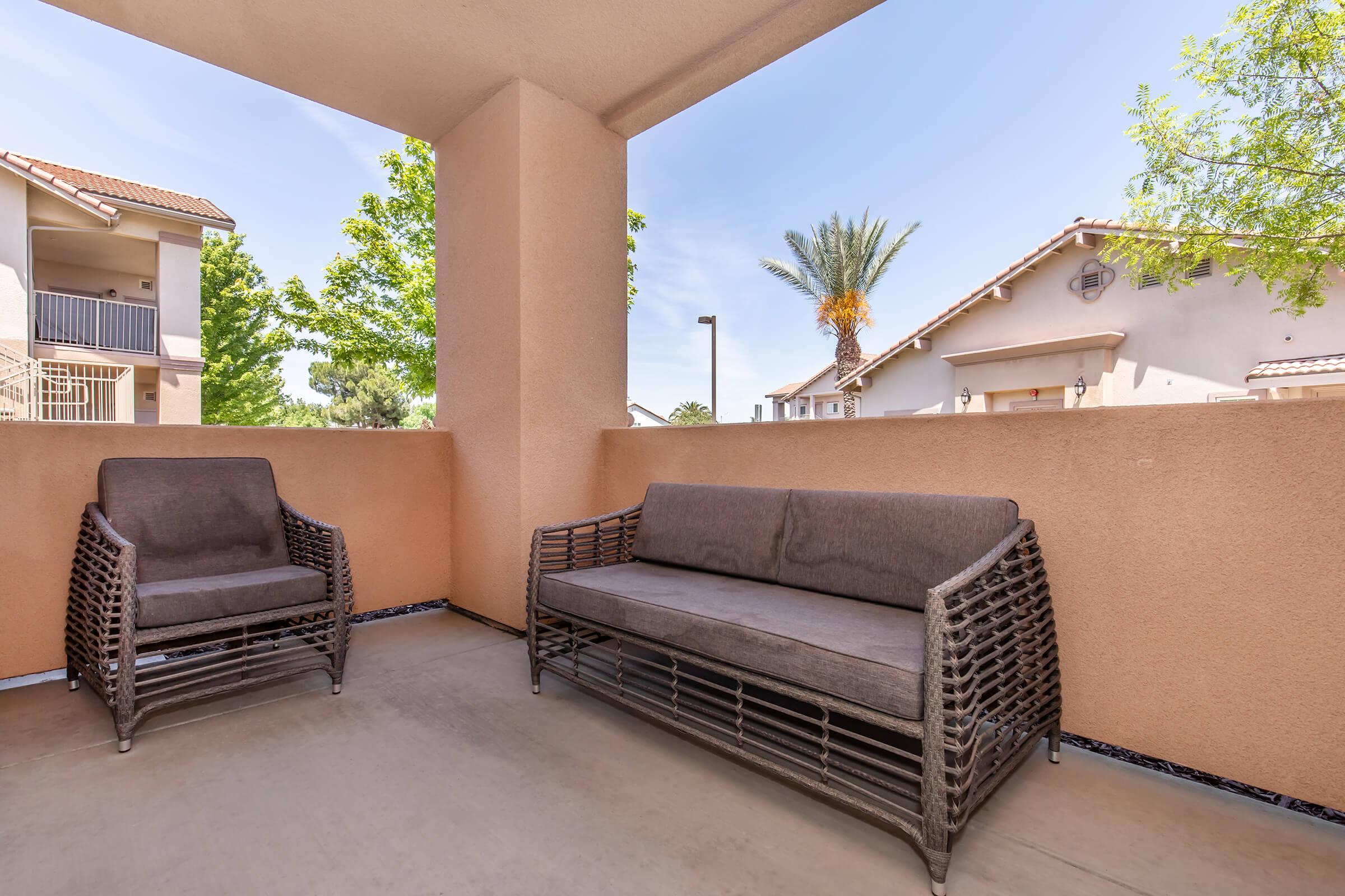 A patio area featuring two wicker chairs and a sofa against a peach-colored wall. In the background, there are residential buildings and palm trees under a blue sky, suggesting a sunny day. The scene creates a relaxed, outdoor living space.