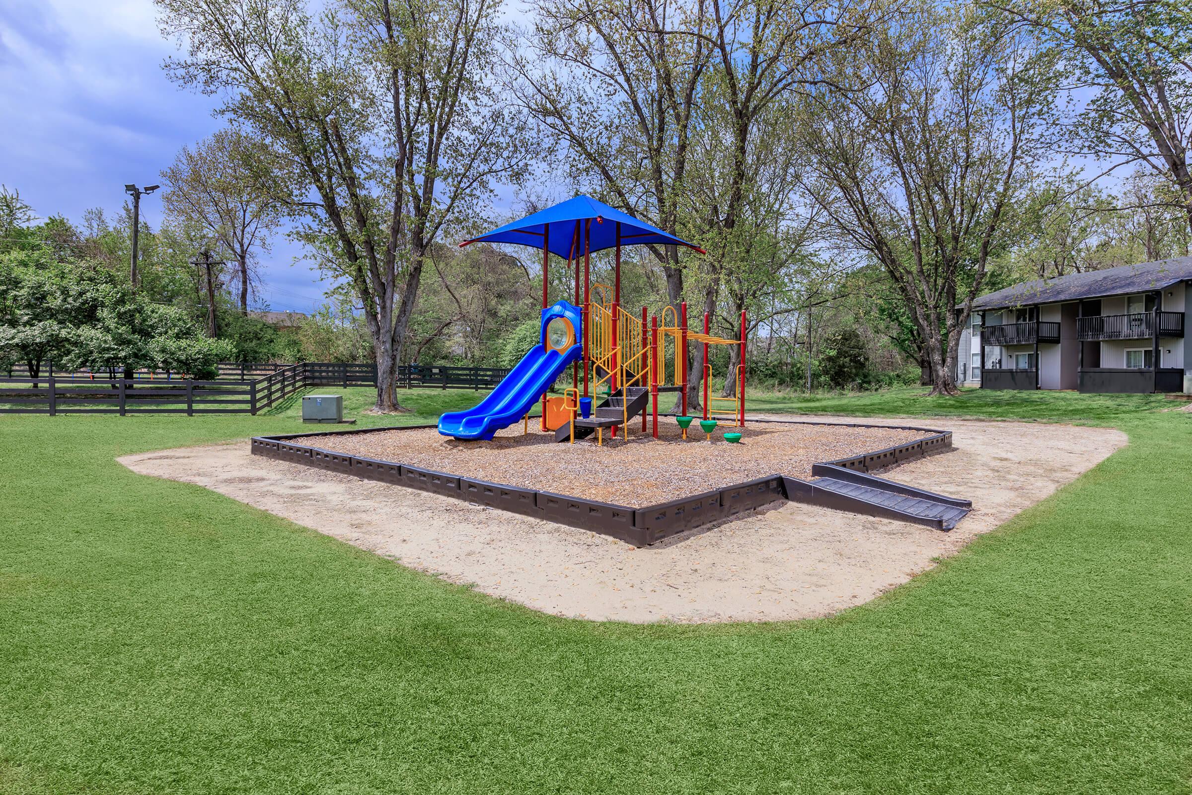 A colorful playground featuring a blue slide, climbing structure, and shaded area under a large blue canopy, all situated on a sandy surface surrounded by green grass and trees. In the background, there are residential buildings and a wooden fence.