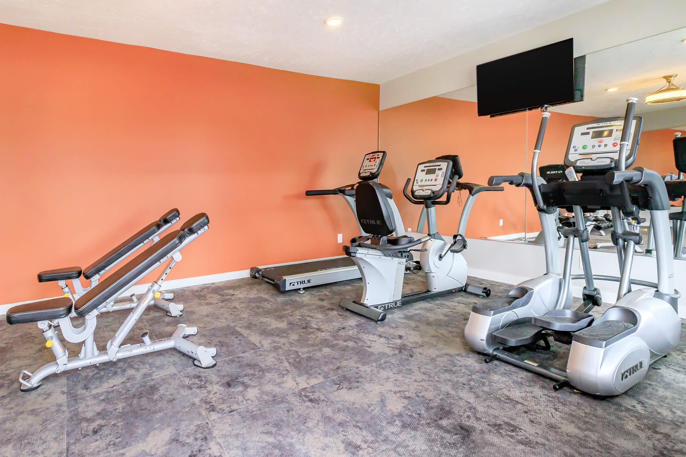 A modern gym area featuring exercise equipment including a bench press, stationary bike, and elliptical machine. The walls are painted orange, and there is a large mirror and a flat-screen TV mounted on the wall, creating a bright and inviting workout space.