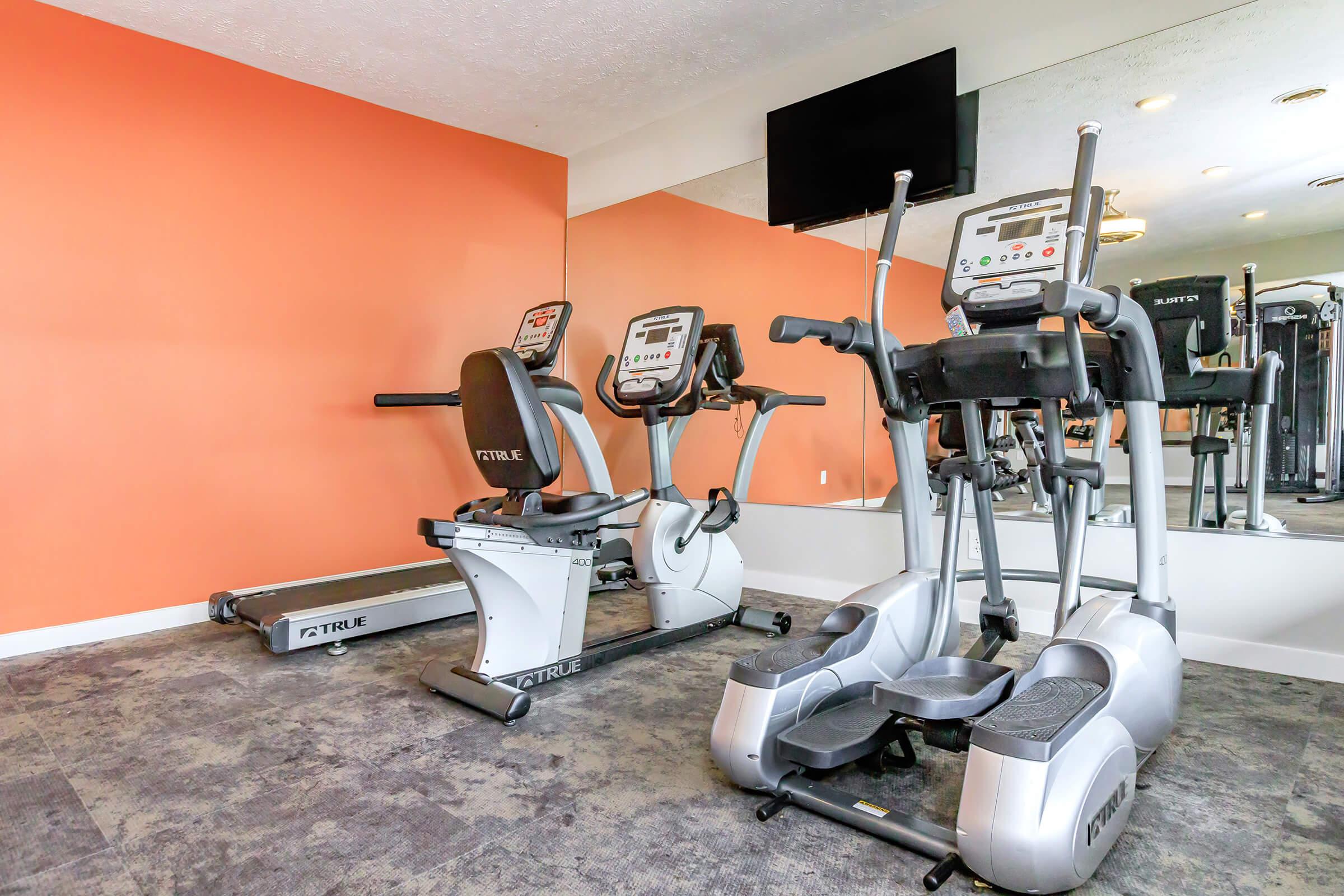 A well-equipped gym interior featuring a treadmill and an elliptical machine against an orange wall. A television is mounted on the wall, and large mirrors reflect the exercise equipment. The floor is covered with gray carpet, creating a modern and inviting workout space.