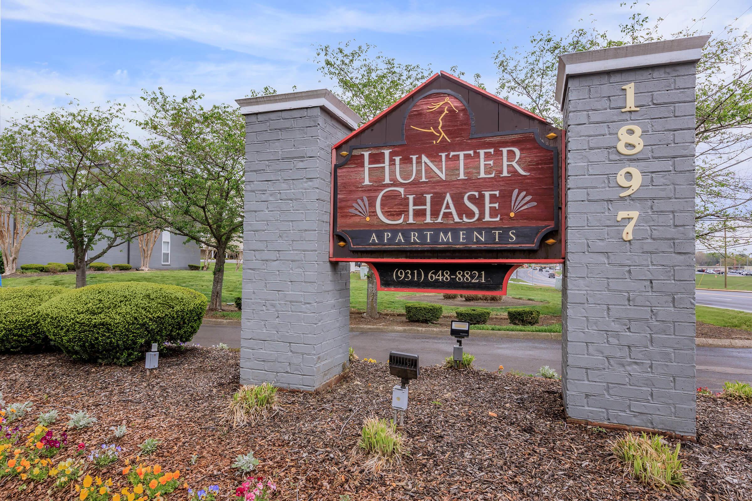 Sign for Hunter Chase Apartments, featuring a wooden design with the name prominently displayed. The structure is flanked by decorative landscaping and greenery, with a stone base that includes the number 1897. Contact phone number (931) 648-8821 is also visible.