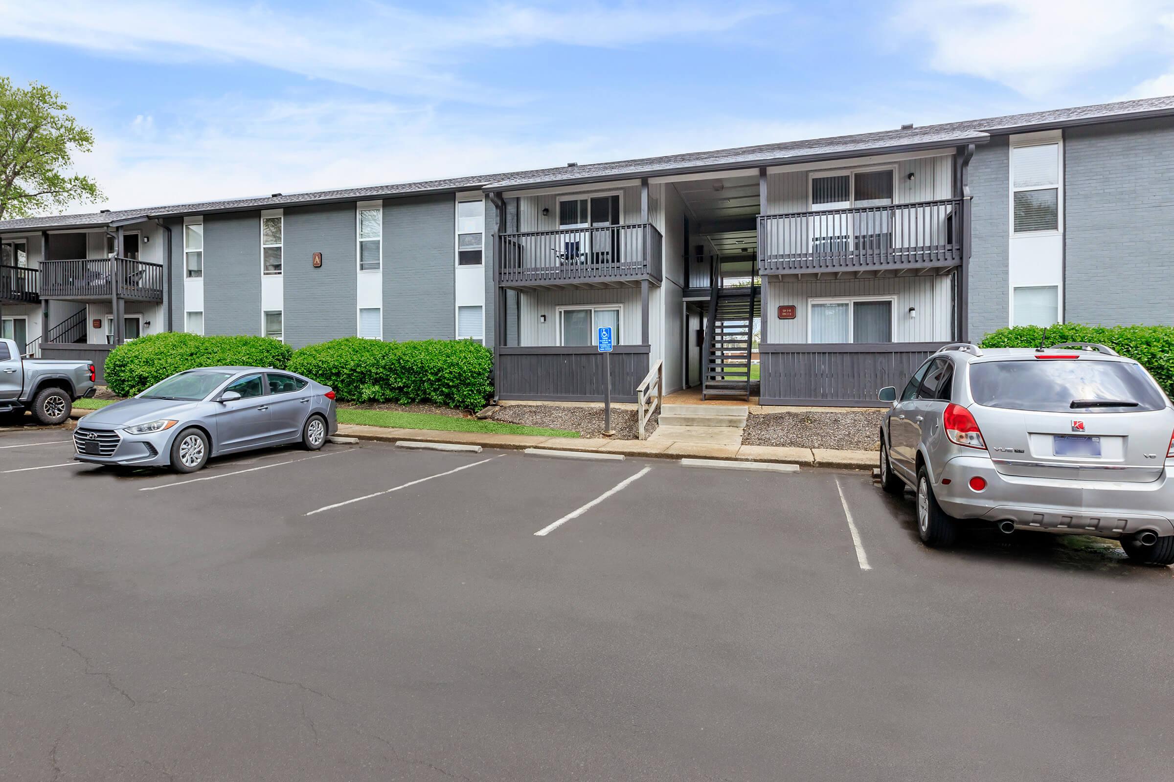 A two-story apartment building with gray exterior, featuring balconies on the upper level. The foreground shows a parking lot with several cars parked. Lush green bushes border the building, and there are visible walkways leading to the front entrances.
