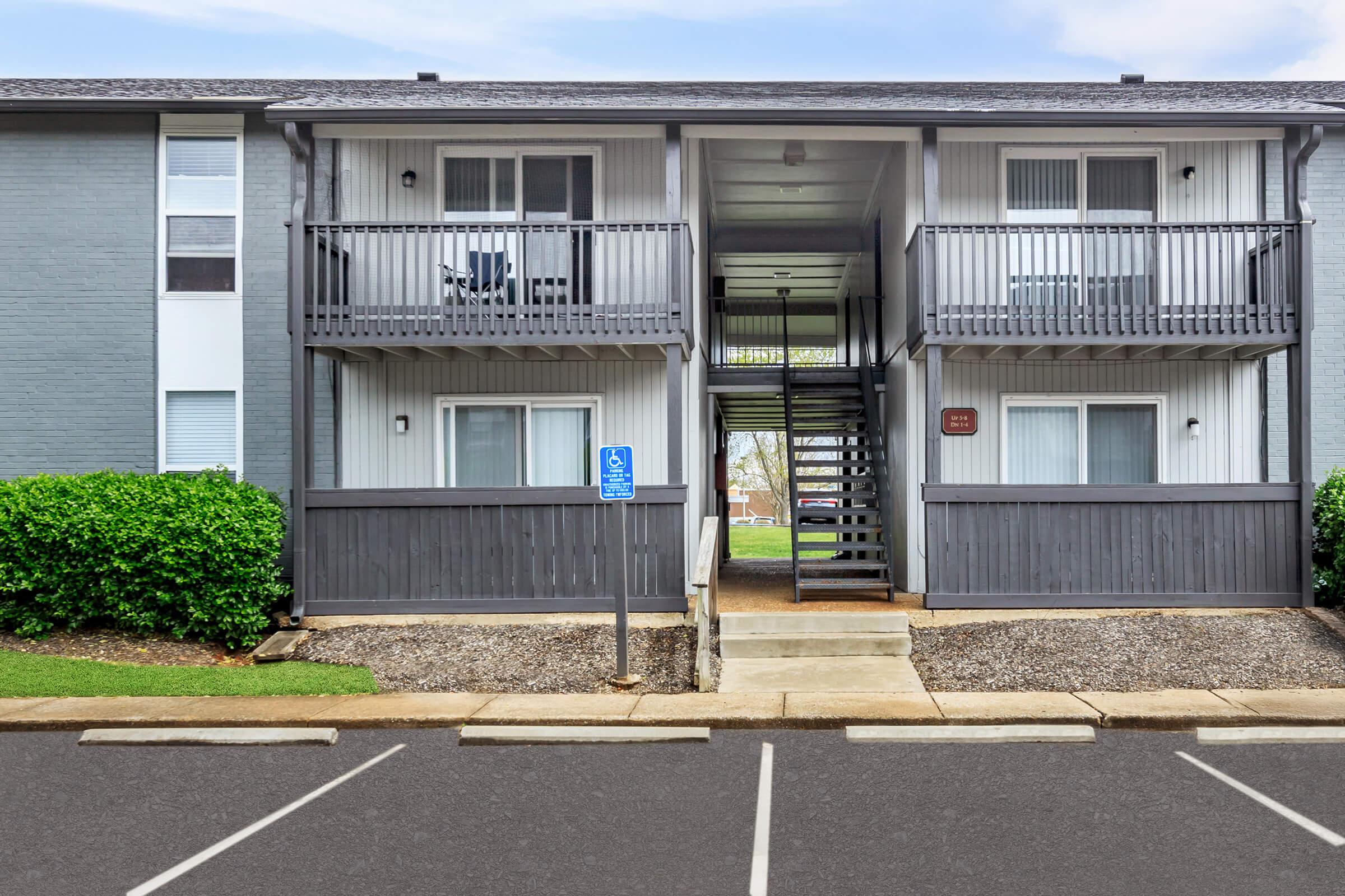Two-story apartment building with a gray exterior, featuring two units on the upper level with balconies. There is an outdoor staircase leading to the second floor. A designated parking space is visible in front of the building, along with landscaped greenery.