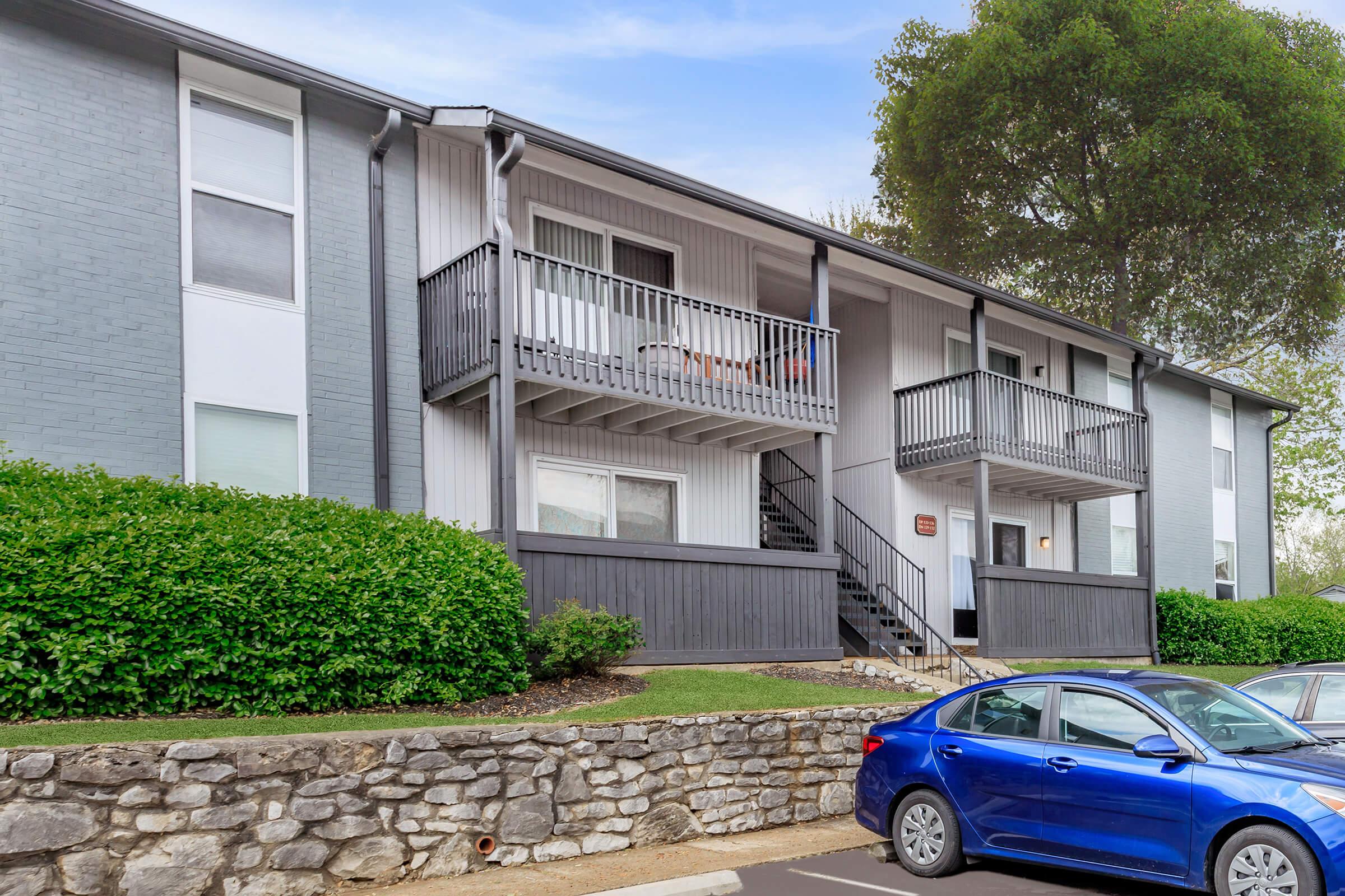 A two-story apartment building featuring balconies with wooden railings. The exterior is a mix of gray and white, with well-maintained landscaping including bushes. A blue car is parked in the foreground, and there are several parked cars visible in the lot.