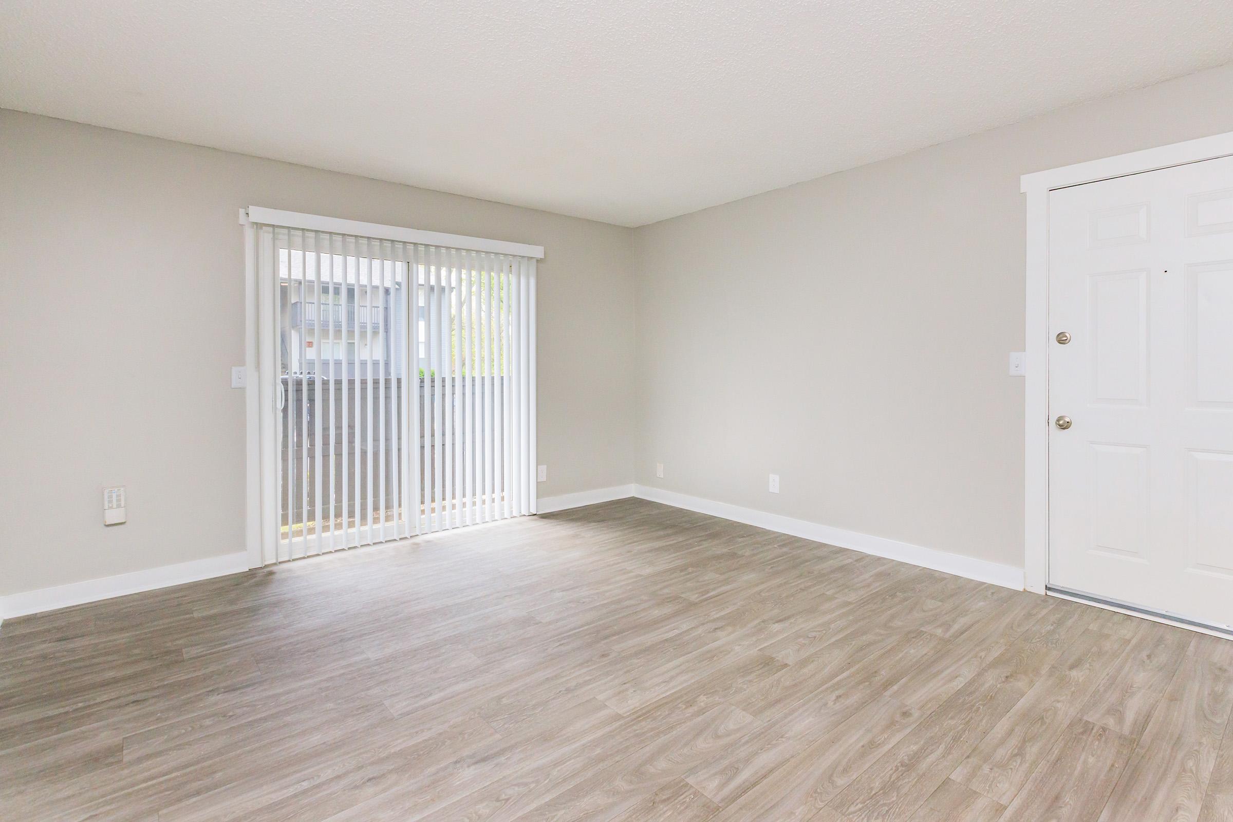 A spacious, empty room with light-colored walls and a wooden floor. On one side, there’s a sliding glass door with white blinds, providing natural light. The entrance features a white door with a doorknob, and the overall ambiance is bright and inviting.