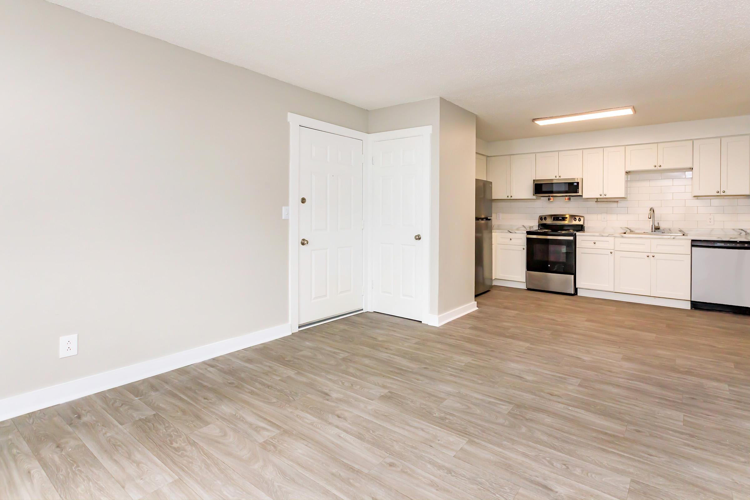 Interior view of a modern, well-lit kitchen and living area. The kitchen features white cabinets, stainless steel appliances, and a light countertop. A door with double panels is visible on the left, with light-colored walls and a laminate floor creating a spacious, inviting atmosphere.