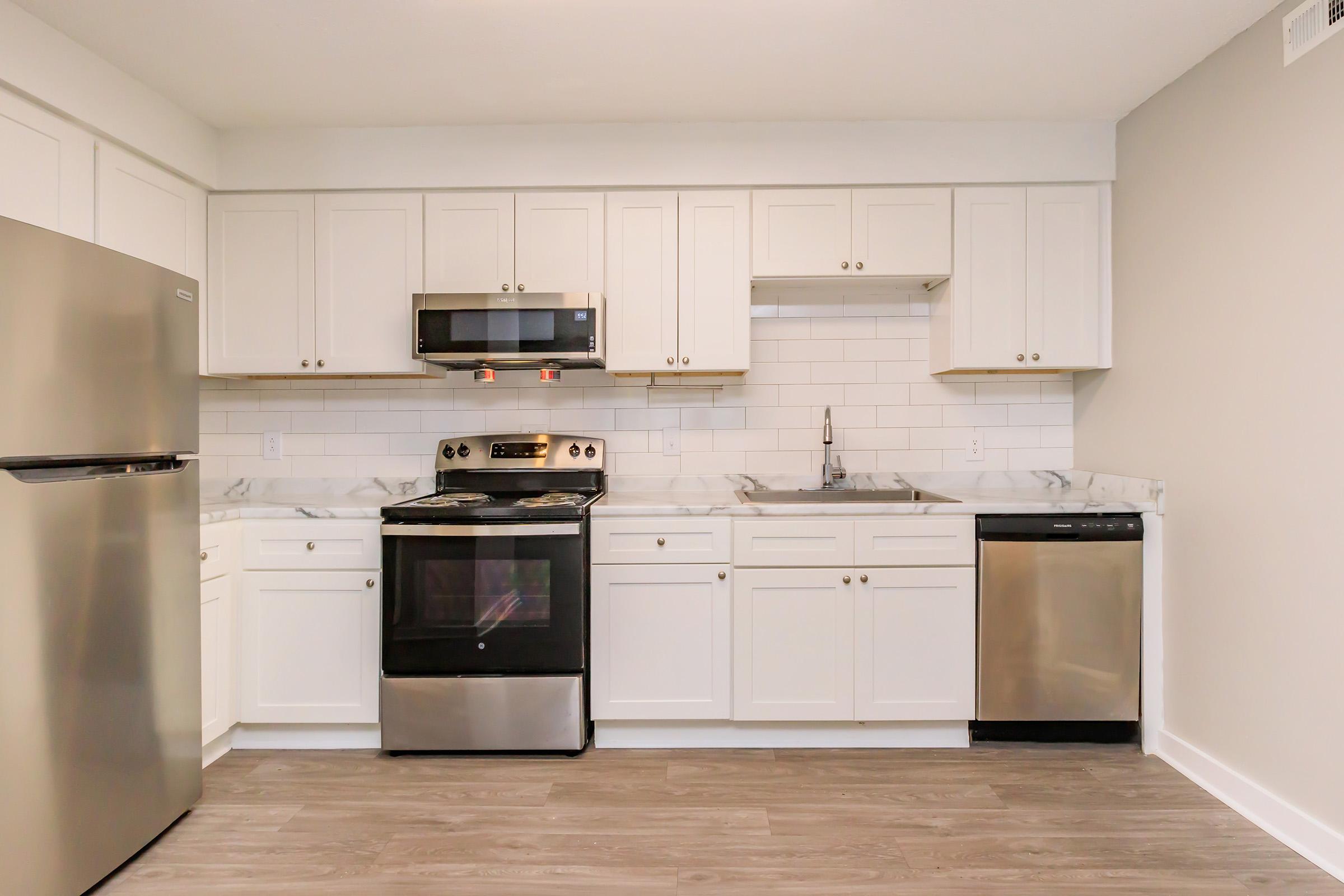 Modern kitchen with white cabinets, stainless steel appliances including a refrigerator, stove, and dishwasher. The countertop features a marble design, and there's a white subway tile backsplash. The floor is a light wood finish, creating a bright and clean aesthetic. Natural light is present from an unseen window.
