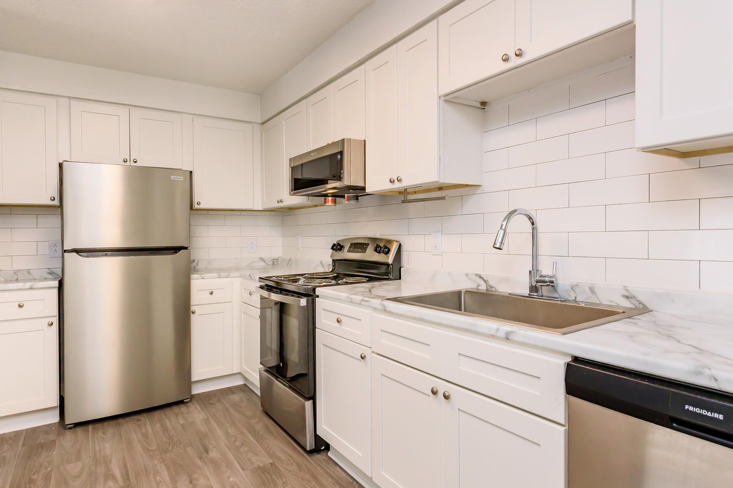 Modern kitchen featuring stainless steel appliances including a refrigerator, microwave, and stove. Light-colored cabinetry and a marble countertop complement the white subway tile backsplash. A stainless steel sink is situated under a window, and the flooring is a light wood finish, creating a bright and inviting space.