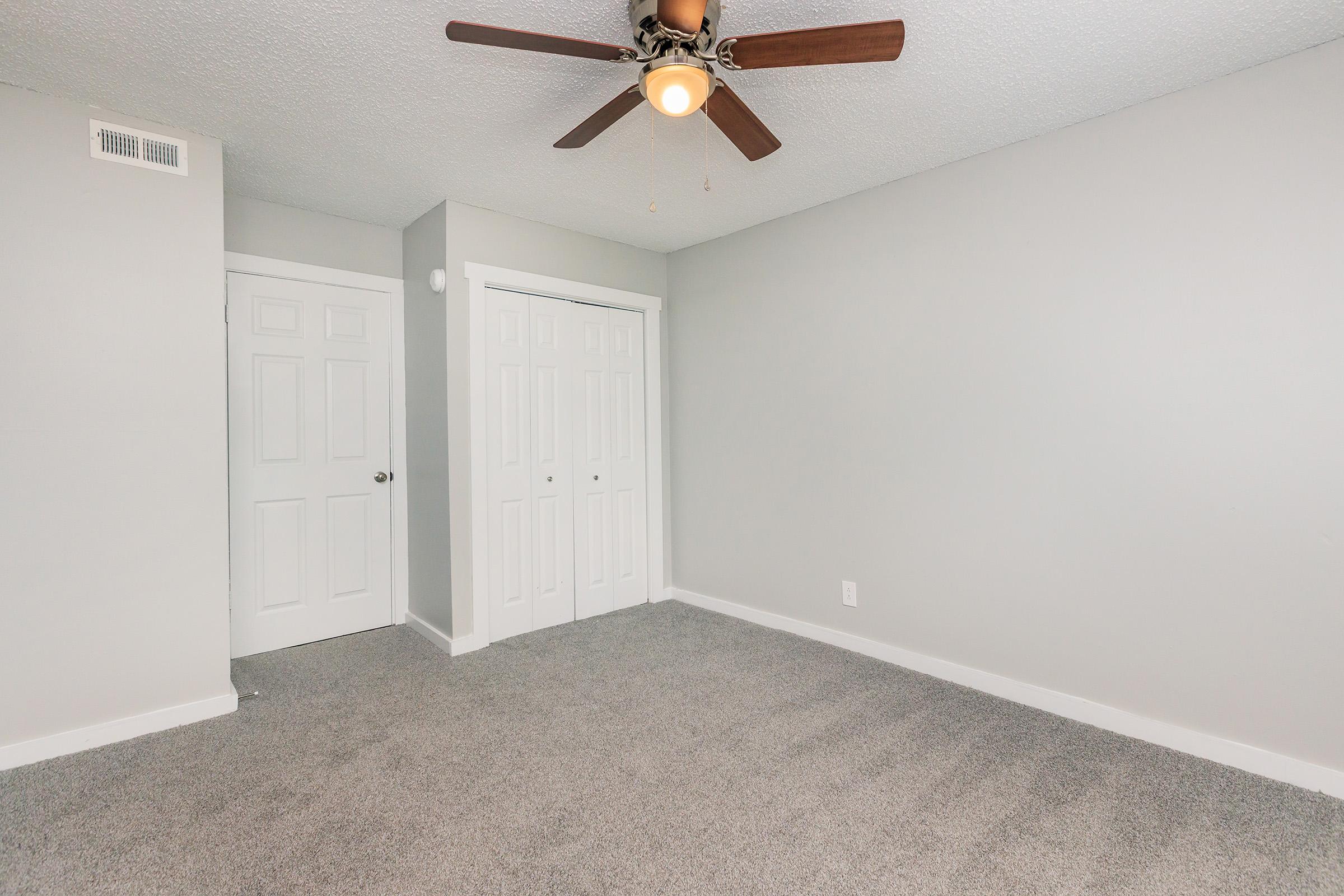 A spacious, empty room with light gray walls and a ceiling fan. The floor is covered with plush gray carpet. There are two white sliding doors on the left, leading to a closet. Natural light is coming in, giving the room a bright and airy feel.