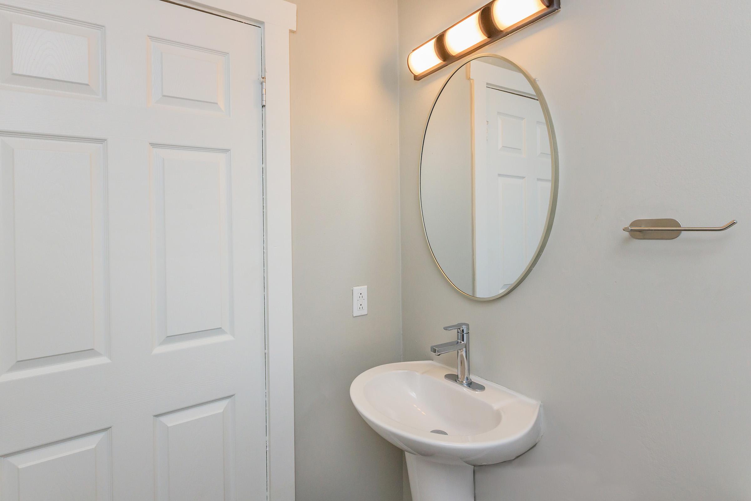 A modern bathroom featuring a white circular mirror above a sleek white sink, with a contemporary faucet. The light fixture above the mirror has three bulbs, providing illumination. A white door is visible on the left side, and a towel bar is mounted on the wall next to the sink. The overall color scheme is light and neutral.