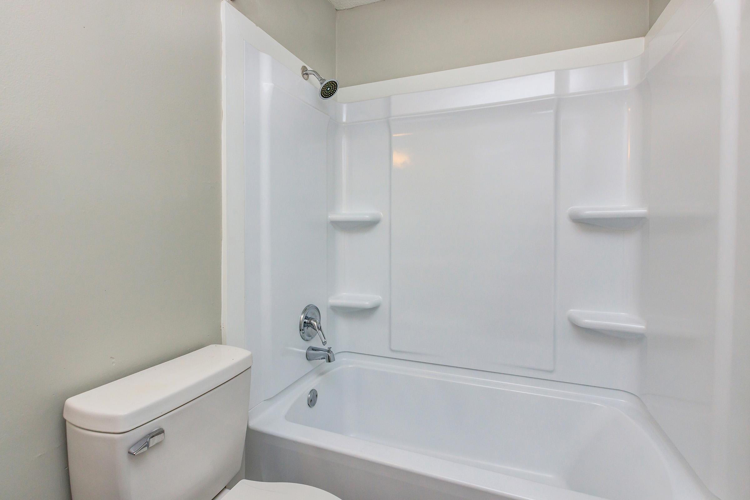 A clean, modern bathroom featuring a white bathtub with an integrated shower, a wall-mounted showerhead, and light-colored walls. The toilet is positioned next to the bathtub, creating a functional and minimalist space. No decorations or personal items are visible, emphasizing simplicity.