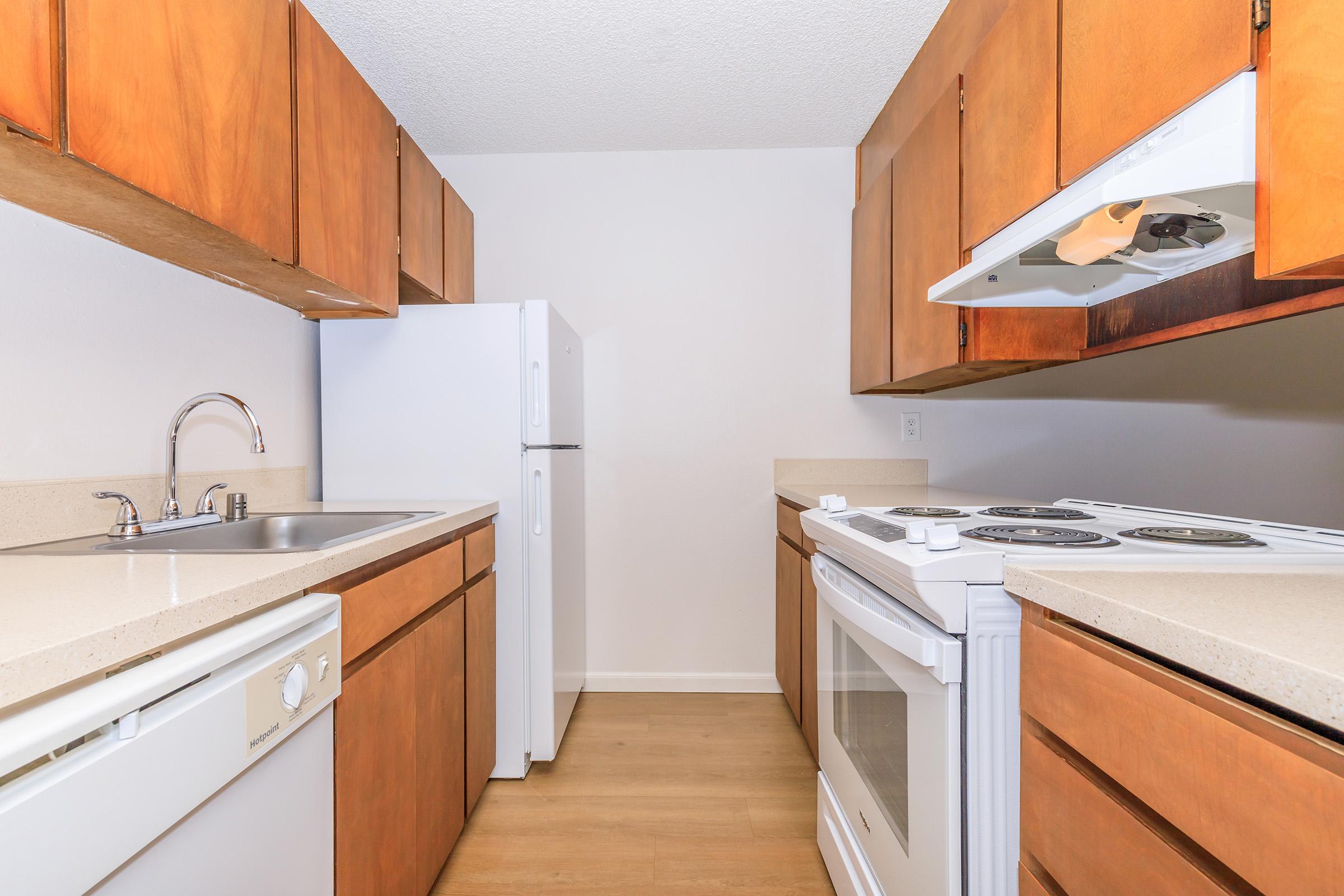 A modern kitchen featuring wooden cabinets, a stainless steel sink, a refrigerator, and a white stove. The countertops are beige, and the flooring is light wood. The kitchen is well-lit and organized, creating an inviting atmosphere.
