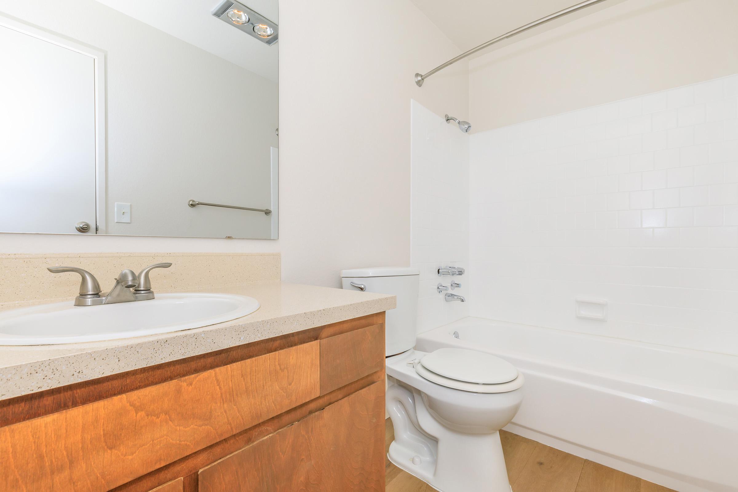 A clean and minimalistic bathroom featuring a light-colored countertop with dual sinks, a wooden cabinet below, a white bathtub with a shower, and a toilet. The walls are light and the flooring is wood, creating a bright and spacious feel.
