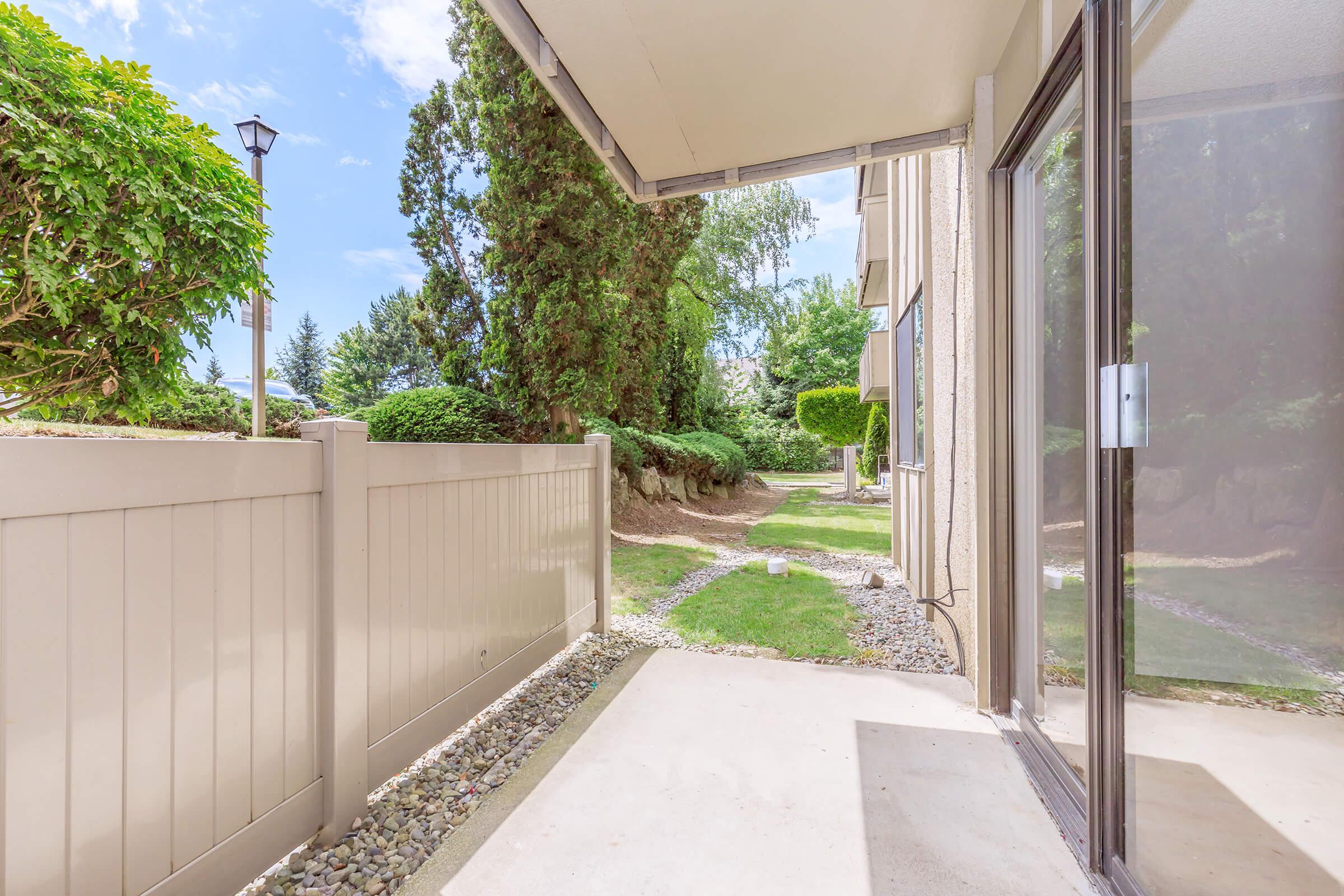 A patio area with a view of a well-maintained garden and trees in the background. The beige fence borders the patio, and the ground is paved with light-colored concrete. Bright blue sky with scattered clouds is visible above. The setting appears peaceful and inviting.