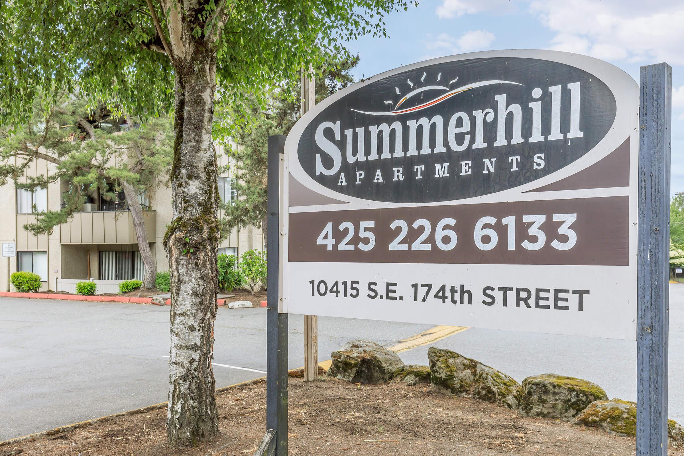 Sign for Summerhill Apartments with contact number 425-226-6133 and address 10415 S.E. 174th Street. The sign is located in front of the apartment complex, surrounded by trees and a parking area.