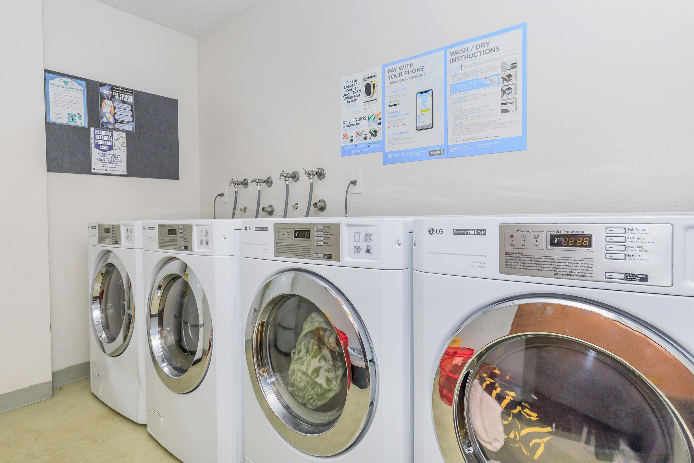 A row of four white front-loading washers and dryers in a laundry area. Each machine features circular glass doors, digital controls, and a sign with washing instructions on the wall above. The floor is light-colored, and there are additional informational posters on the wall.